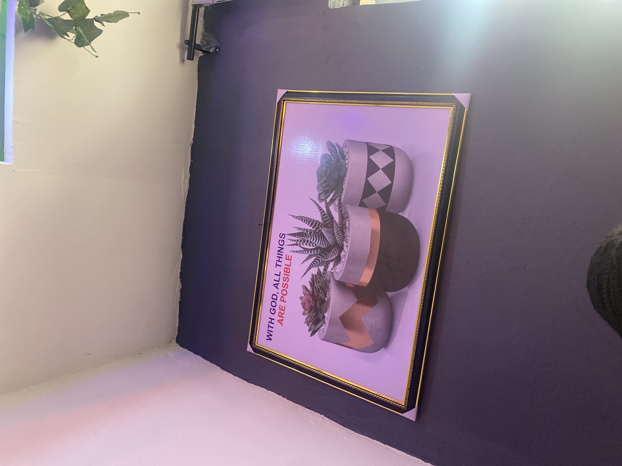 An framed print featuring three succulent plants in decorative pots is displayed on a dark purple wall. The print has a gold and black border with a Greek key pattern. The text "WITH GOD, ALL THINGS ARE POSSIBLE" is printed in red and purple above the succulents. The lighting in the room is dim and has a purple hue, possibly from ambient lighting or a colored light source. To the right of the print, a black bracket or fixture is mounted on the wall. The ceiling is white. The image appears to have been taken in a residential or commercial indoor space. The location within the city of Rafin Par, Nigeria is not discernible from the provided image.