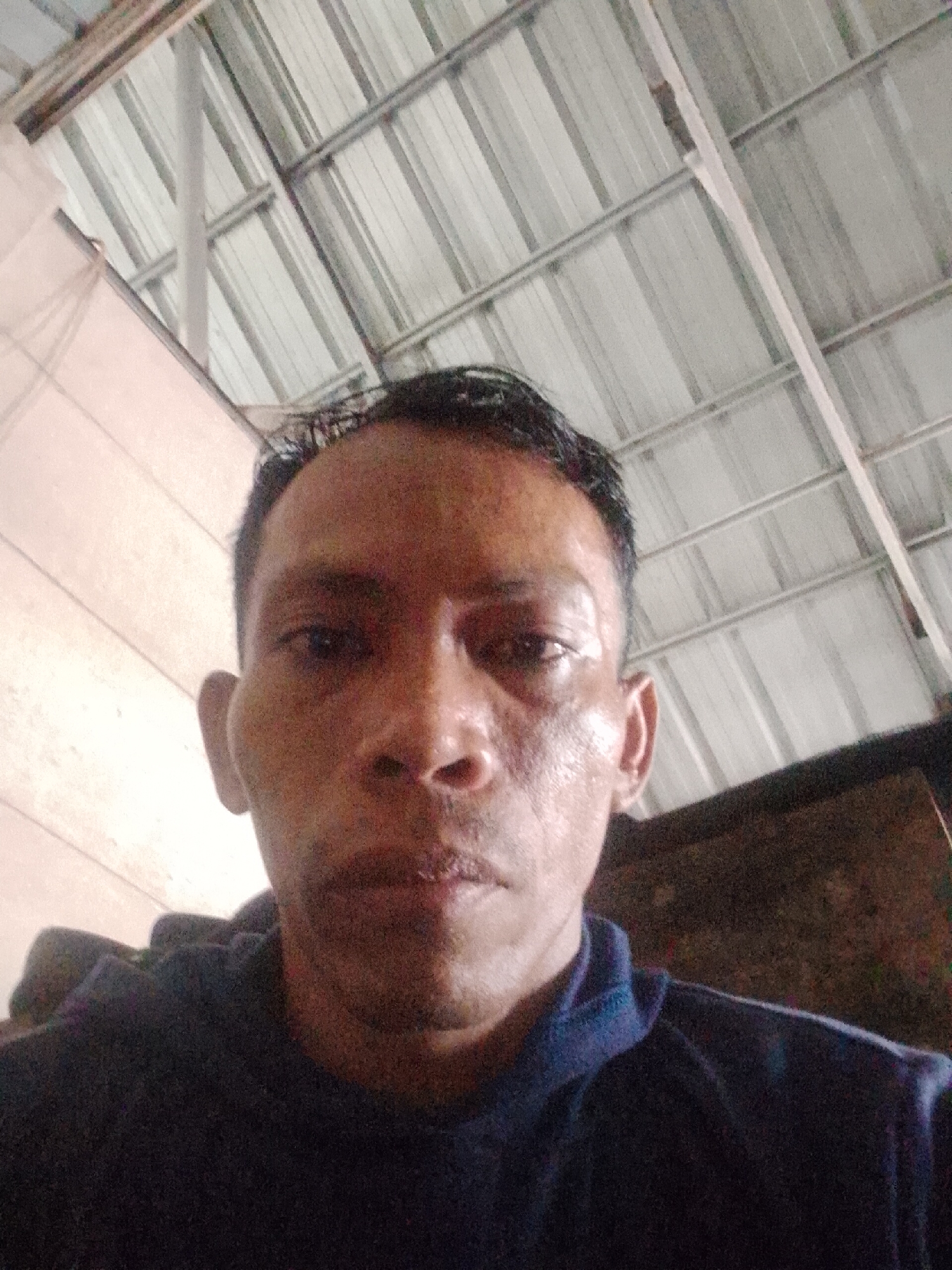 The image shows a close-up, low-angle shot of a man. He appears to be indoors, as a corrugated metal roof structure is visible overhead. The man is wearing a dark blue top. The lighting is dim, and the background is somewhat blurred. Bukit Ketok, Indonesia is provided as the location.