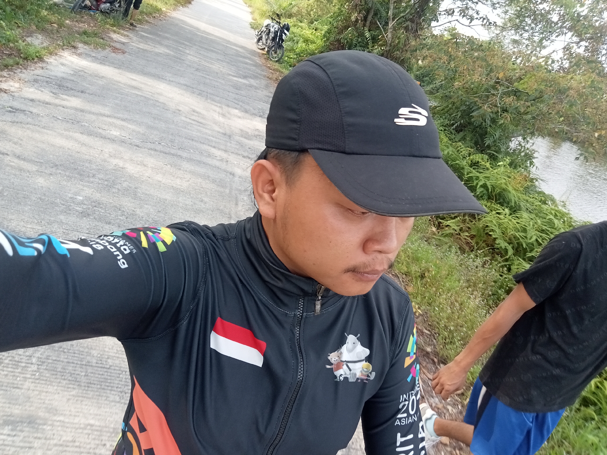 The image shows a selfie taken by a person wearing a black cap and a black jacket with colorful graphics and the Indonesian flag. The person is standing on a paved road, possibly in Kualu, Indonesia, with a body of water and lush greenery on the right side of the frame. In the background, two motorcycles are parked on the side of the road. Another person is partially visible on the right, wearing a black t-shirt and blue shorts. The overall lighting suggests it is daytime, possibly morning or late afternoon.