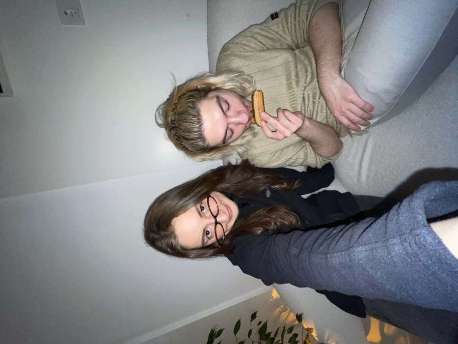 Two individuals are captured from an elevated angle. One fair-haired person in a beige sweater and light gray pants appears to be eating a pastry. Beside them is someone with brown hair and glasses, wearing a black jacket and dark gray pants. They are both reclining on a white couch. A plant is partially visible in the bottom left corner of the frame. A wall with an electrical outlet is in the background. The photo has a somewhat dim and casual aesthetic.