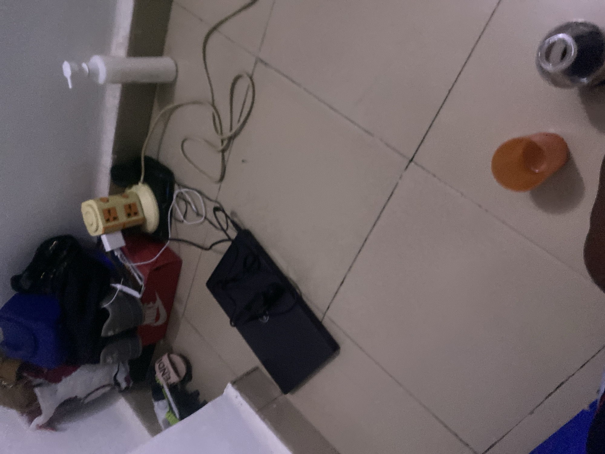 The image depicts a cluttered corner of a room, likely a bedroom or storage area, with objects scattered on a tiled floor. In the background, near a wall, are several items including a white dispenser bottle, a yellow and orange electric fan with an exposed power cord, and various bags and shoes. A red Nike shoe box is visible, containing gray shoes. A black laptop is placed open on the floor in the mid-ground, with its charging cable connected. A pair of headphones rests on the laptop. In the foreground, there is an orange-colored object, possibly a cup or lid, and a dark, metallic object with a reflective surface. The floor is made of beige square tiles. The overall impression is one of disarray. The location is identified as Howayi, Nigeria.