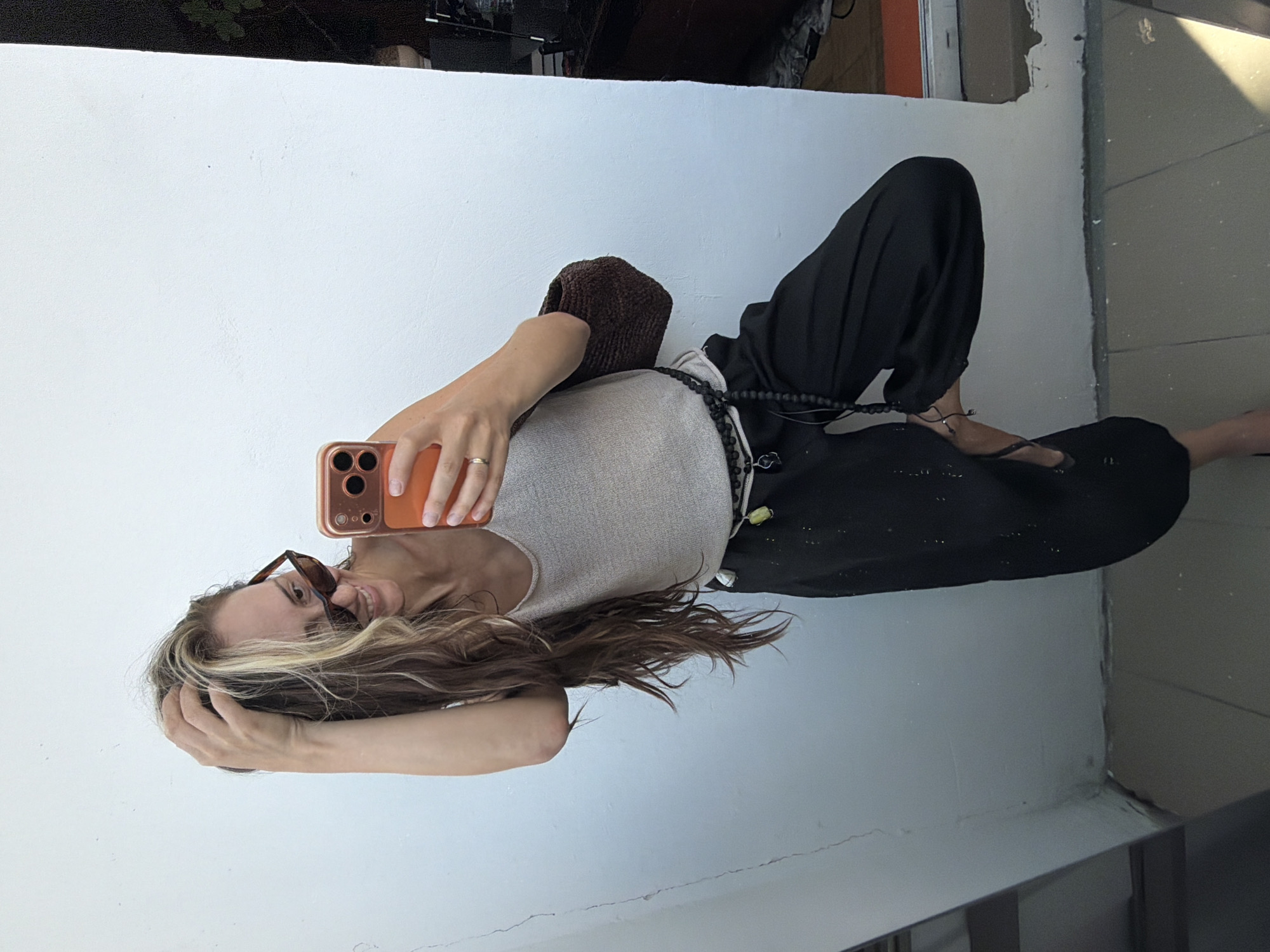 A young Caucasian woman with long, blonde and brown hair, wearing sunglasses, a beige tank top, black pants, and a beaded belt, is taking a selfie with an orange iPhone in her right hand. She is standing on a tiled floor in front of a white wall. Her left hand is raised and touching her hair. A brown knitted bag is slung over her left shoulder. The environment suggests an outdoor urban setting, possibly a balcony or a patio, with a wall and floor visible. There is no discernible indication of the location being Moscow, Russia.