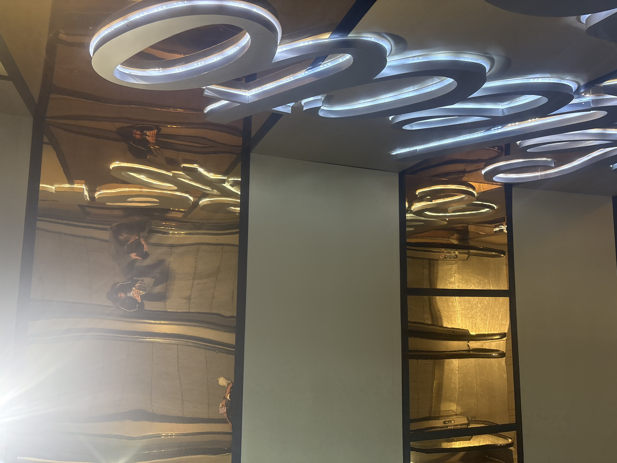 The image shows a ceiling with a modern lighting fixture in an indoor space, likely a commercial building or hotel in Mexico City, Mexico. The lighting fixture appears to be made of several white, illuminated rings and curved shapes. The ceiling is reflective, creating mirrored images of people walking below. The lighting fixture is mounted onto a grid-like structure with dark framing. The walls are off-white, and a window or display case with shelves is visible on the right. People are faintly reflected walking under the ceiling on the left side of the image.