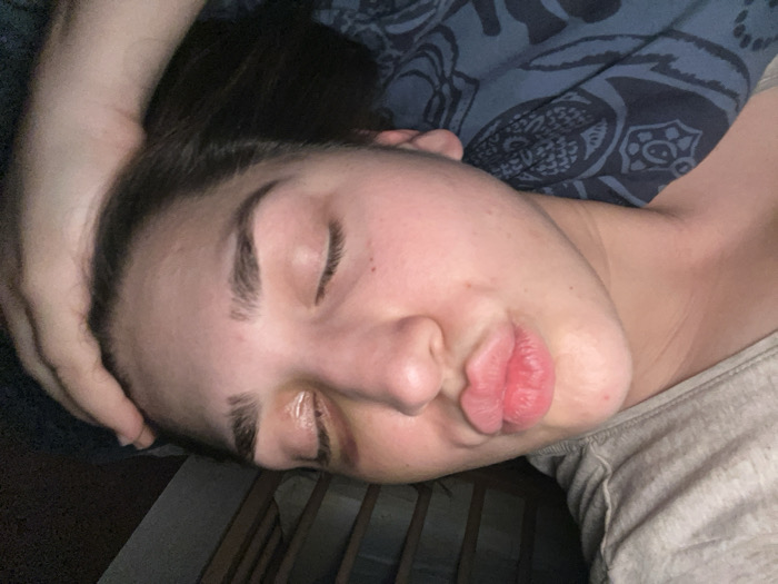 A woman with closed eyes and pursed lips rests her head on someone's arm. She has dark hair, prominent eyebrows, and fair skin. A blue patterned fabric is near her head. She is wearing a light-colored t-shirt. The background appears dark and slightly out of focus, suggesting an indoor setting.