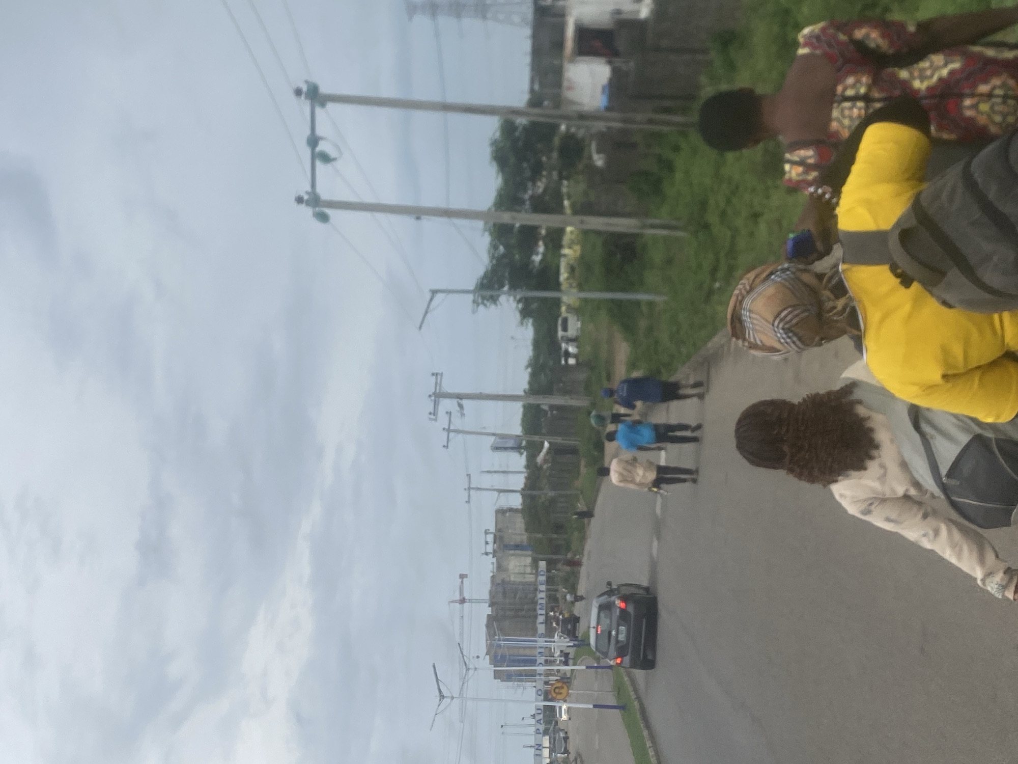 The image depicts an outdoor street scene in Lugbe, Nigeria, under an overcast sky, suggesting daytime. The perspective is from the side of a paved road that extends into the distance.
In the foreground, several people are walking along the roadside. Closest to the viewer, a person is seen wearing a bright yellow top with a dark backpack. Adjacent to them, another individual with curly brown hair wears a light-colored top and dark lower attire. Further along, a person in a brown and white checkered pattern top with a blue cap, and another in a vibrant red, yellow, and black patterned dress, are visible. More pedestrians, some dressed in blue shirts, can be observed further down the road.
A black car is driving on the road in the mid-ground. The area is characterized by numerous utility poles and overhead power lines running alongside the road. Lush green grassy areas and trees line the right side of the road. In the background, several buildings, some appearing to be under construction with visible cranes, are present. A large high-tension electricity pylon stands prominently in the far background against the cloudy sky.
A sign visible in the mid-ground along the road displays partial text, which reads "IN PAU O" and "S LIMITED", possibly indicating a business or development name like "IN PAULO HOMES LIMITED". The scene captures everyday activity in a bustling, semi-urban environment.