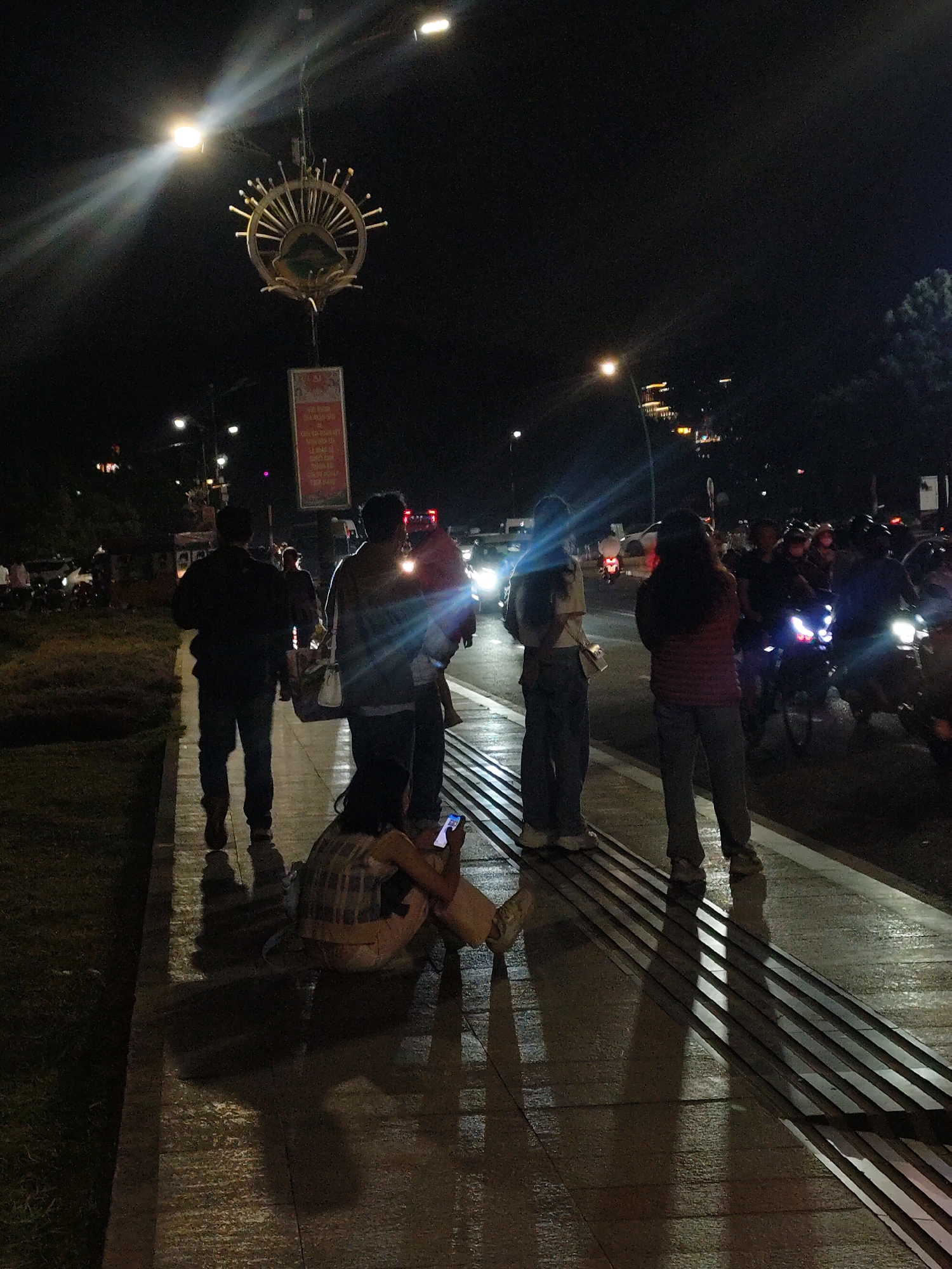 This image depicts a nighttime scene in Da Lat, Vietnam, likely on a street or pedestrian walkway. In the foreground, a person is sitting on the ground, illuminated by the light of their mobile phone. A few feet away, four individuals stand on a sidewalk, their silhouettes cast by the streetlights and passing vehicle headlights. The sidewalk features tactile paving. Behind them, a street is active with motorcycle traffic and other vehicles, their lights creating streaks of illumination. A large decorative structure hangs from a lamppost overhead, with a banner displayed below it. The background is dark, with some distant lights suggesting buildings and trees. The overall atmosphere is urban and nocturnal.