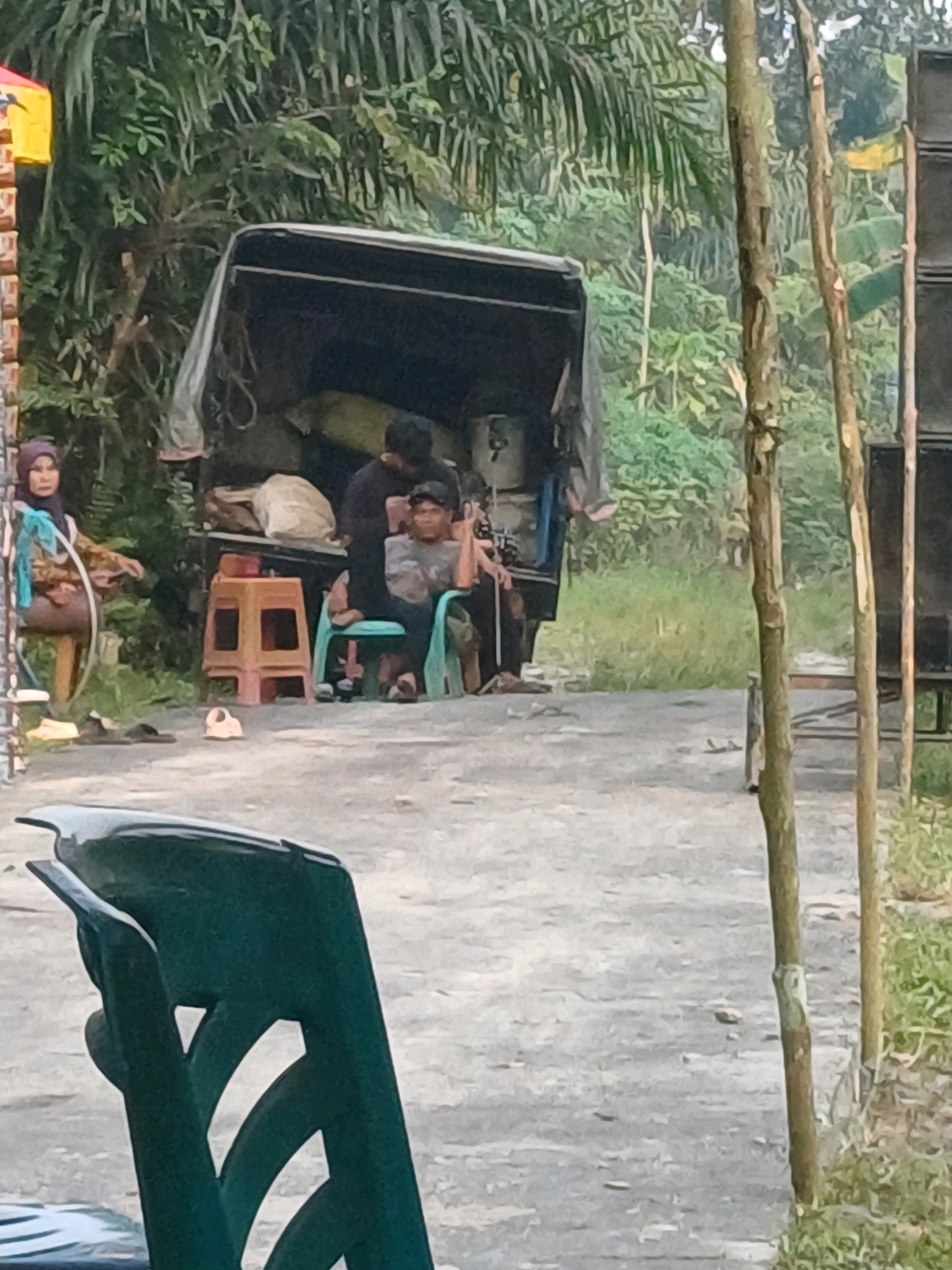 The image depicts an outdoor scene in Dumai City, Indonesia, with a focus on a pickup truck and several individuals. In the foreground, a green chair is prominently featured. Behind the chair, a dirt or gravel path leads towards the truck. The truck itself is partially loaded with goods.
To the left of the truck, a woman wearing a hijab and a patterned top is seated. She appears to be observing the scene. Two men are positioned near the back of the truck; one is partially visible, and the other is sitting in a green chair, looking towards the viewer. A small, orange stool is located between the woman and the man in the green chair.
The background consists of lush green vegetation, including trees and foliage, suggesting a rural or semi-rural setting within Dumai City. Two vertical poles or tree trunks are visible on the right side of the image, with what appears to be a speaker system mounted on the rightmost one. The overall lighting suggests it is daytime.