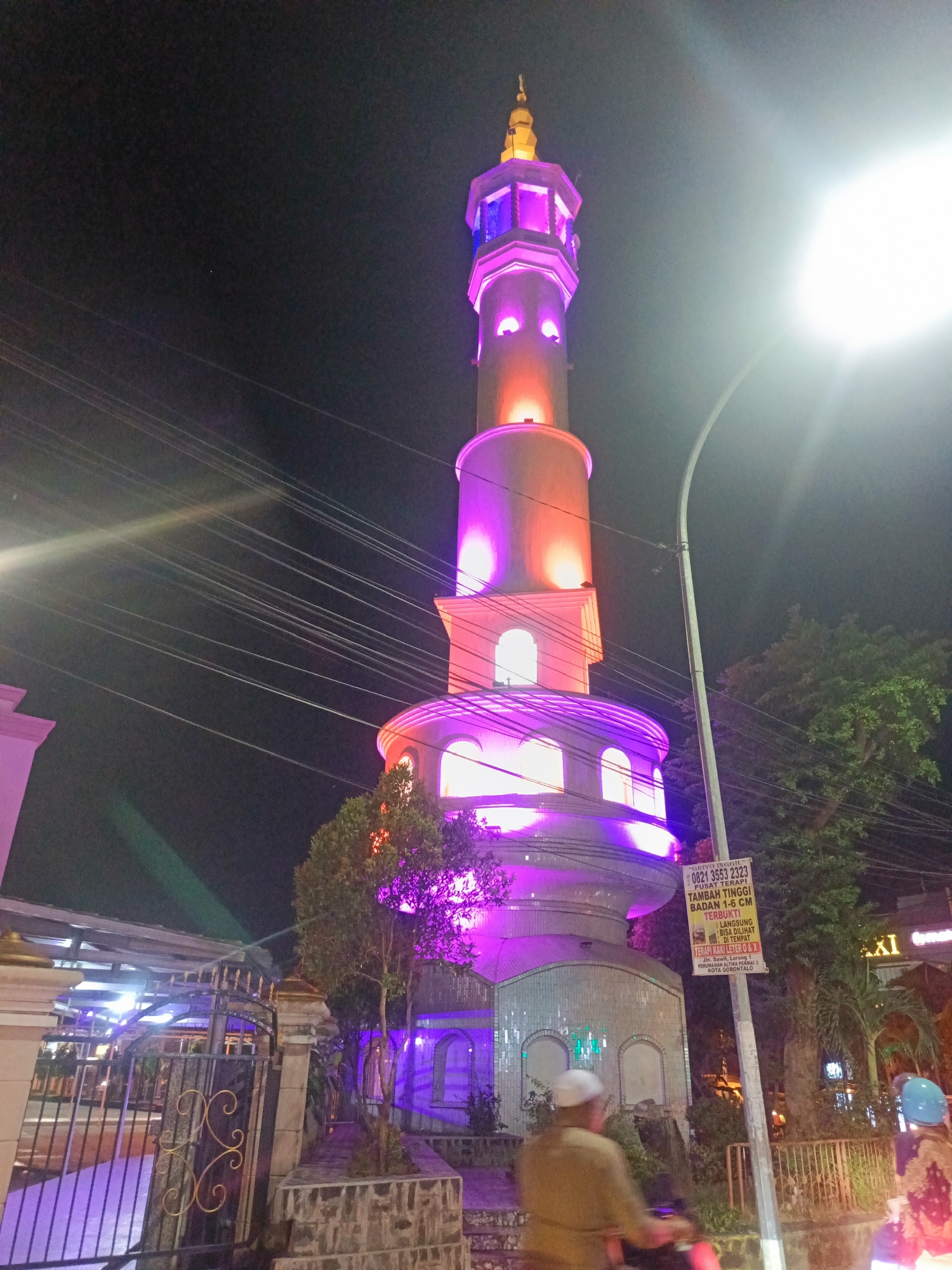 At night in Gorontalo, Indonesia, a brightly lit minaret stands tall, its upper sections illuminated in shades of pink and orange, with purple lights at its base. The minaret has a rounded base, a cylindrical shaft, and a tiered upper section topped with a golden spire. Arched windows are visible on the minaret's structure. In the foreground, a black metal gate with ornate gold scrolling is partially visible to the left, leading to a walkway. A lush green tree grows near the minaret's base. To the right of the minaret, a lamppost displays a sign with Indonesian text and a phone number. Beyond the minaret, trees and the illuminated windows of buildings can be seen. The sky is dark, with power lines stretching across it. The scene suggests a public space or the vicinity of a mosque, with the lighting designed for visual appeal at night.