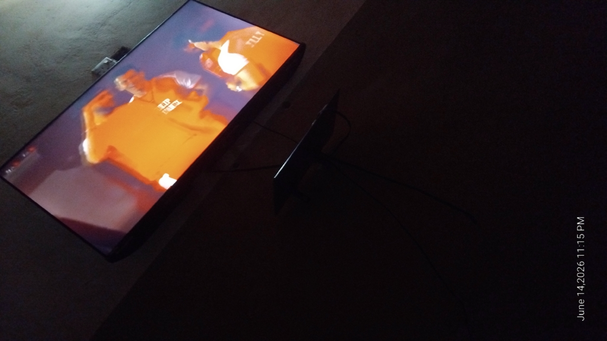A television screen displays a dynamic image of two individuals in what appears to be a sporting event or performance. The screen is angled upwards and to the right, with the surrounding environment largely in shadow. The individuals on screen are wearing brightly colored, predominantly orange, clothing. One individual is in the foreground, their body contorted in motion, with visible text on their attire that reads "SLIP STREAM". The other individual is behind them, also in motion, with visible text on their clothing that reads "TILLY". The lower portion of the image shows a dark, unidentifiable object mounted below the television, with wires extending from it into the darkness. A timestamp in the bottom left corner indicates "June 14, 2026 11:15 PM". The overall scene suggests an indoor setting at night, possibly a living room or entertainment space, where a vibrant broadcast is being viewed in low light conditions.