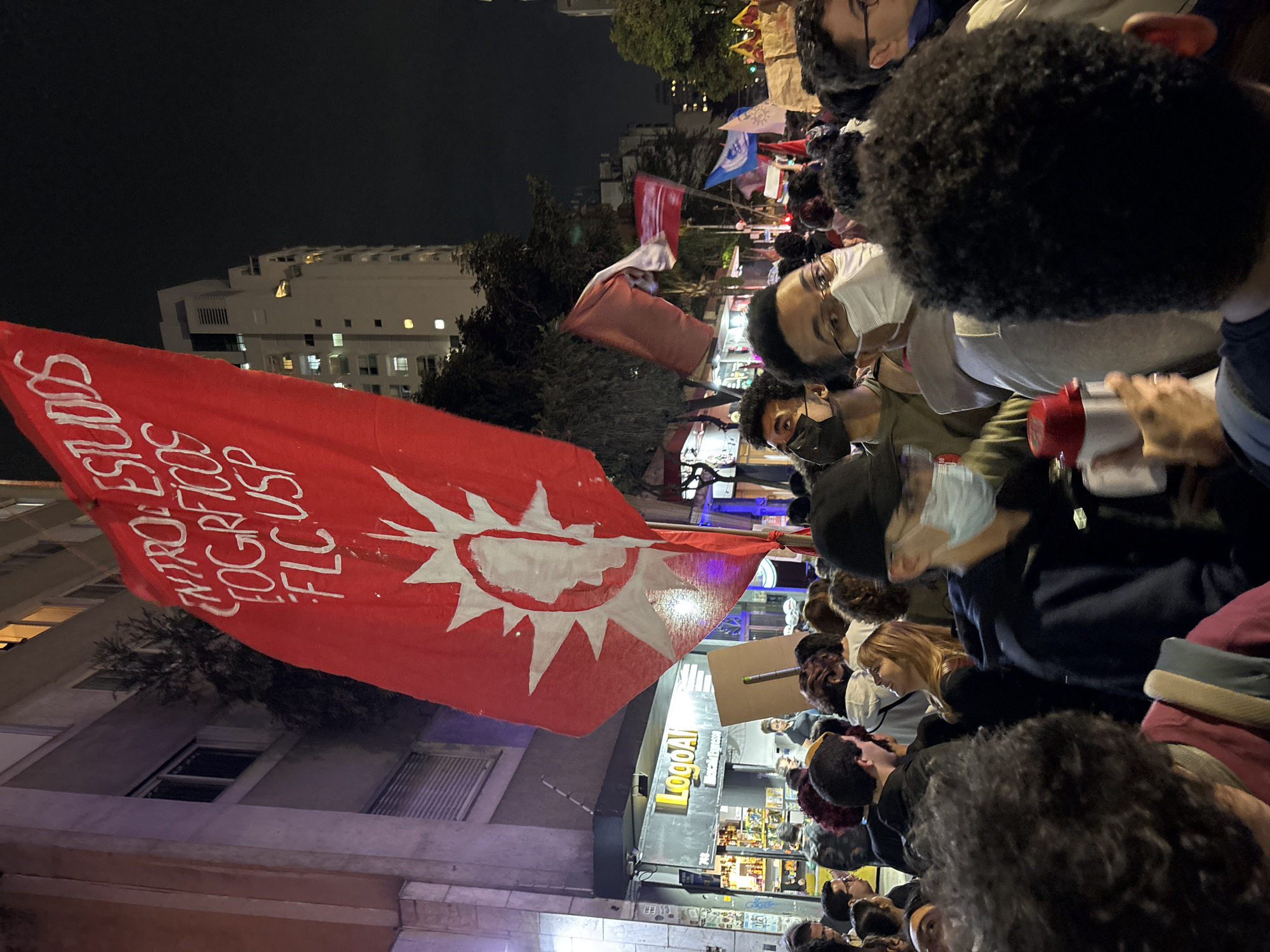 A nighttime outdoor scene captures a large crowd, likely a protest or demonstration, in what appears to be an urban area of São Paulo, Brazil. The focal point is a red flag emblazoned with white text and a sun-like symbol, hoisted above the crowd. The text, visible in Portuguese, partially reads "CONTROLE ES... GEOGRAFICOS... FLC USP," indicating a possible connection to the University of São Paulo's geography department.
The crowd is dense, consisting of individuals of varied ages and ethnicities. Several people are wearing face masks, suggesting a recent or ongoing public health concern. At least one individual is using a megaphone.
In the background, buildings are illuminated, and the presence of street-level businesses is noticeable; a sign with "LogoAI" is partially visible. The overall atmosphere suggests a politically charged gathering in a vibrant urban setting.