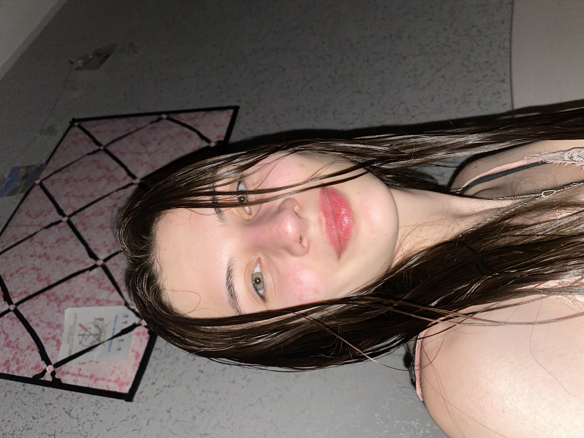 The image, rotated 90 degrees counter-clockwise, captures a young woman lying down indoors in dim lighting. She has long, dark brown hair that appears wet or damp, spread around her head and shoulders. She looks directly at the viewer with a neutral expression, light-colored eyes, and lips tinted pink. Her fair skin shows a slight flush on her nose and cheeks.
She is wearing a light pink or beige top with lace trim and a visible black strap, along with a thin silver chain necklace.
To her left, a notable object is visible: a pink surface adorned with a black grid pattern, possibly a decorative mat or a small kite. Affixed to this object is a white label containing text and symbols. While the text is partially obscured and blurry, it appears to be in a non-Latin script, consistent with Cyrillic, given the location context of Votkinsk, Russia. One identifiable symbol on the label resembles a "no entry" or "do not touch" sign. The surrounding surfaces are dark and textured. The low light conditions suggest the photograph was taken in the evening or night.