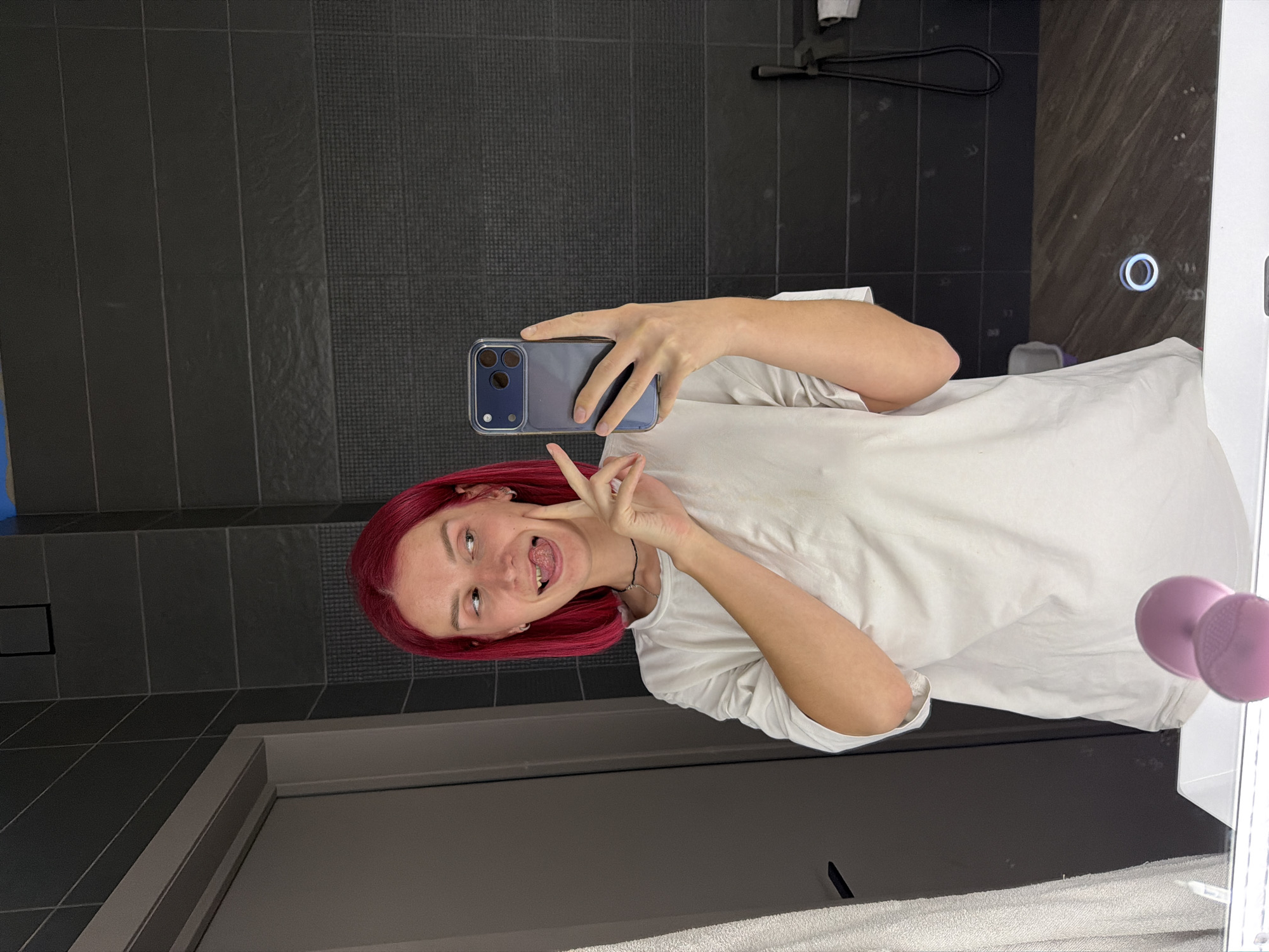 An individual with bright, reddish-pink hair is captured in a mirror selfie, holding a smartphone with a clear case. They are wearing a white t-shirt and a dark, thin necklace. The person is making a playful expression, sticking out their tongue and forming a "peace" sign with their left hand.
The setting appears to be a modern bathroom. The background features dark grey or black wall tiles, some with a smooth finish and others with a textured pattern. A black towel bar or similar fixture is visible on the wall to the left of the person's reflection. On the right side of the mirror, two pink silicone facial scrubbers are suction-cupped. Below the mirror, there's a dark vanity or counter surface. A white, circular illuminated button or switch is visible on the mirror's surface, common in modern bathroom mirrors. A white towel or fabric is visible along the bottom edge of the mirror reflection. No discernible text is visible in the image. The overall mood conveyed is casual and lighthearted.
