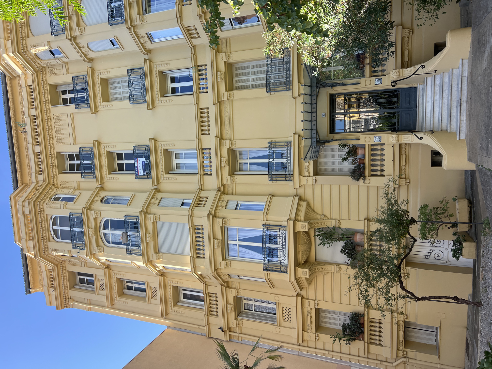 The image shows the facade of a pale yellow, multi-story building with a clear blue sky overhead. The building exhibits ornate architectural details, including decorative moldings, balconies with wrought-iron railings, and arched windows. Several windows have shutters, some of which are open. Potted plants, including cacti and other greenery, are visible on some of the balconies. A "À VENDRE" (For Sale) sign is attached to a balcony railing on the third floor. The ground floor features a dark gray double door entrance with glass panes, approached by a wide set of marble stairs with black metal railings. The number "97" is displayed above the entrance. Trees and foliage are present in front of the building, with a palm tree visible on the left. The scene appears to be a street in Nice, France, as indicated by the architectural style and signage.