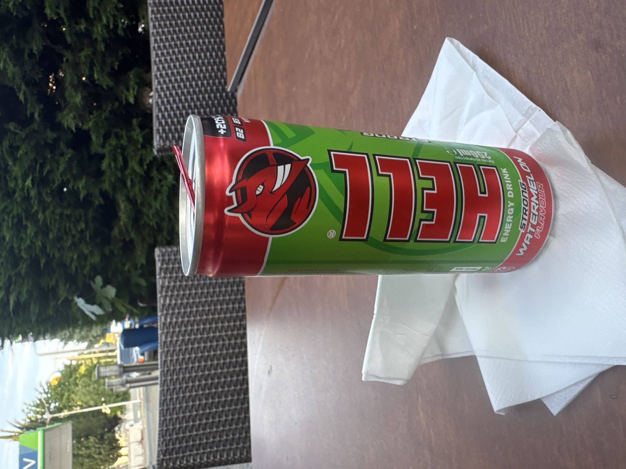 The image shows a green and red "Hell" brand energy drink can, watermelon flavor, sitting on a table with a white napkin. The background shows a street scene with trees and buildings, suggesting an outdoor cafe or restaurant. The location is Stara Zagora, Bulgaria. There are no people visible in the main focus of the image. The energy drink can is the central object.