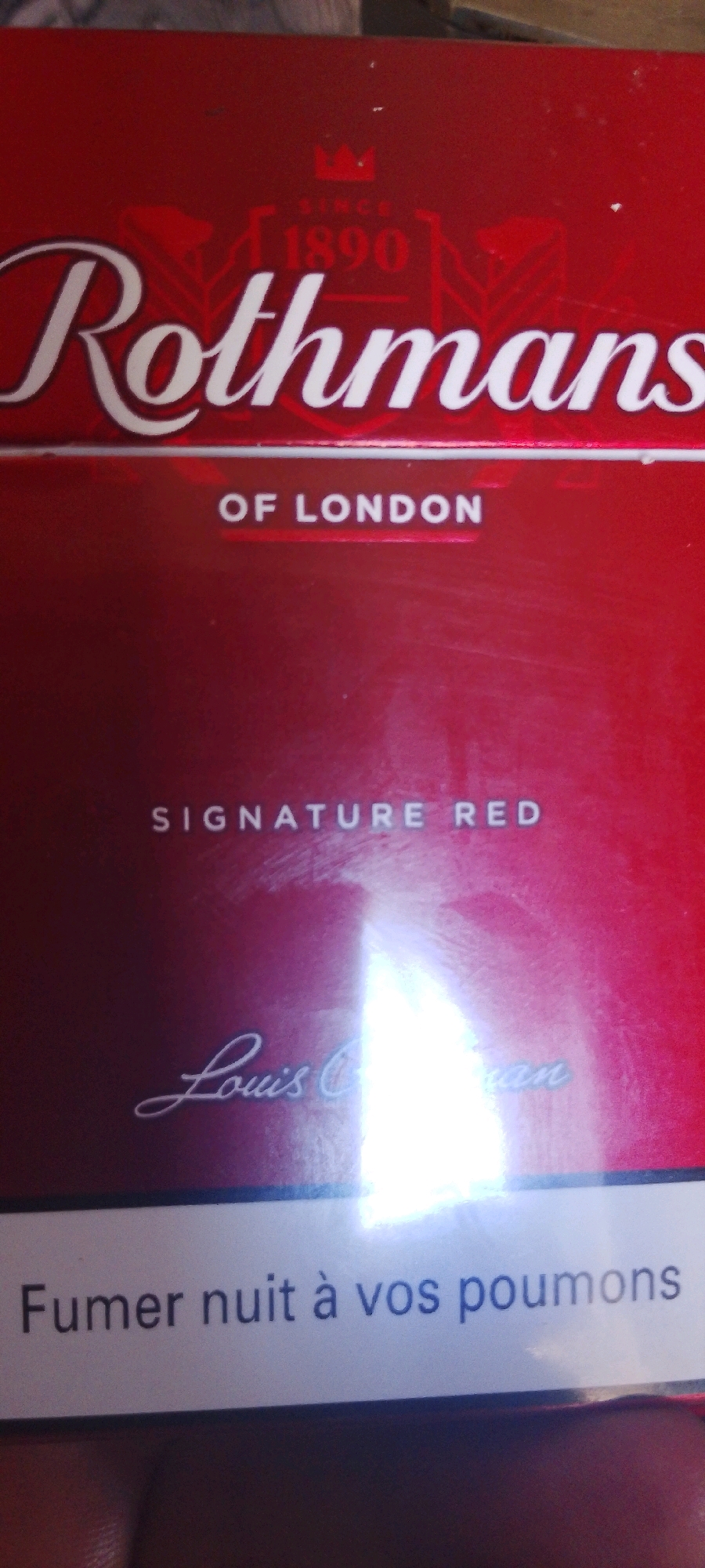 The image shows a close-up of a red cigarette pack with white lettering. The brand name "Rothmans" is prominently displayed in a large, stylized font. Below this, it says "OF LONDON". Further down, "SIGNATURE RED" is written, followed by a signature that appears to read "Louis C. Berman". The bottom of the pack features a white band with black text that reads "Fumer nuit à vos poumons", which translates to "Smoking is harmful to your lungs". The lighting in the image is harsh, creating a bright glare in the center, obscuring some details. The background is dark and out of focus. The image is cropped tightly, showing only the product itself. There are no visible elements related to the city of Moukoudi Ahmed, Algeria.