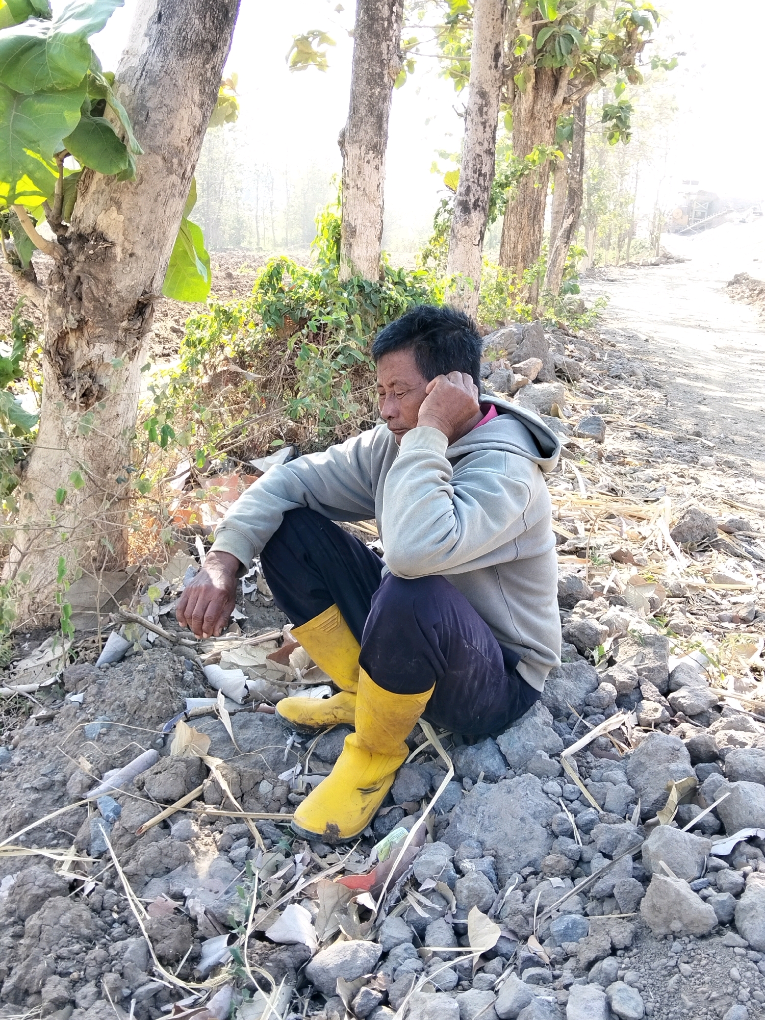 An adult male is sitting on rocky, disturbed ground next to a dirt road. He is wearing a light gray hooded sweatshirt, dark pants, and bright yellow boots. The man is leaning his head on his hand, with his eyes closed, appearing to be resting or contemplating. He is positioned under a large tree with sparse green leaves. The environment is outdoors and appears to be a rural or undeveloped area, possibly on the outskirts of the city of Bujel, Indonesia, given the context. The ground is covered in rocks, dry leaves, and debris. A dirt road with some construction equipment visible in the distance stretches away from the man. The lighting suggests it is daytime, with a bright sky and sunlight casting shadows.