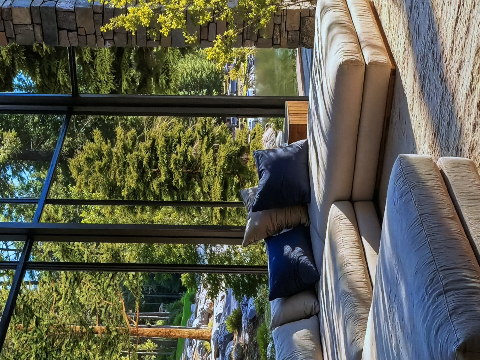 This image captures a serene outdoor living area with a panoramic view of nature. The scene is brightly illuminated by natural sunlight, creating distinct shadows across the furniture.
On the left, large, dark-framed window panels extend, offering an expansive vista of the outdoors. Beyond the glass, a dense forest of lush green trees with vibrant foliage dominates the view. In the mid-ground, a tranquil body of water, likely a lake or pond, is visible, reflecting the bright sky. A section of a stone wall or column is present in the upper left of the frame, bordering the window.
To the right, a comfortable, light-colored outdoor sectional sofa is positioned, featuring multiple thick cushions. Several throw pillows, in shades of dark blue and tan or light brown, are arranged on the sofa. Pronounced, elongated shadows cast by the sun fall across the sofa cushions and a textured, light-colored wall or surface behind the seating. A small wooden side table is partially visible next to the sofa.
The time of day appears to be sunny, either morning or late afternoon, given the clear skies and sharp shadows. No people are present, implying a peaceful moment of relaxation. The overall ambiance suggests tranquility and an intimate connection with the natural environment. There is no visible text in the image. The location context for this scene is Sofiivska Borshchahivka, Ukraine.
