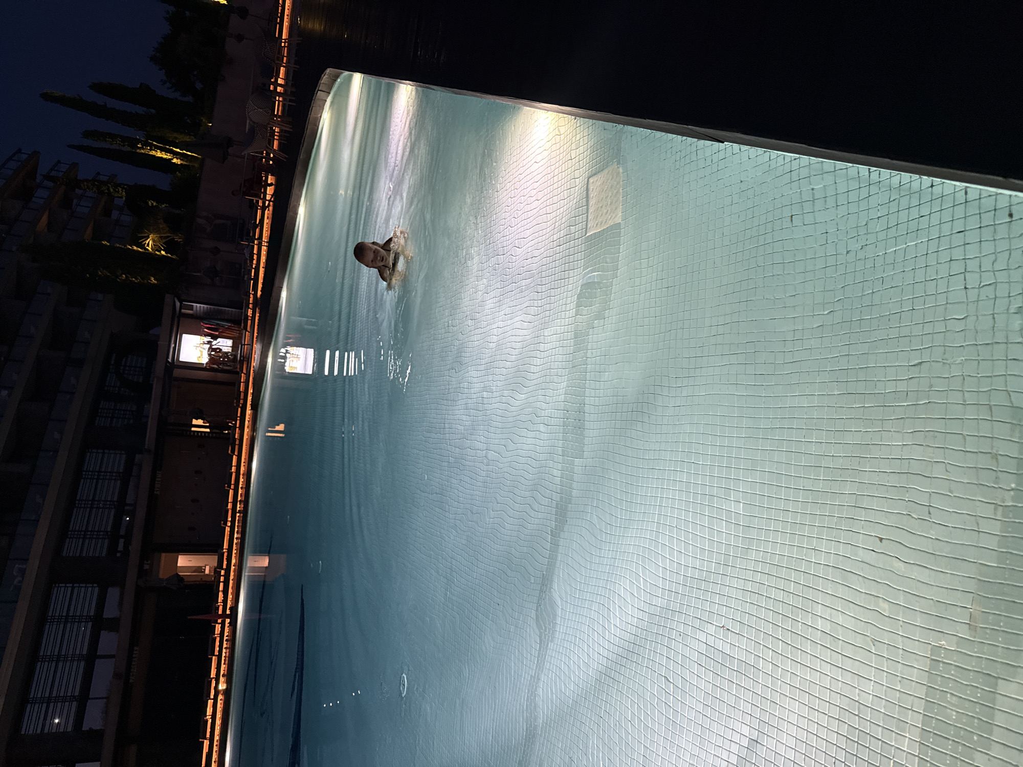 In an outdoor swimming pool at night, a young child with blonde hair is swimming. The pool is illuminated by underwater lights, creating a grid-like pattern on the tiled bottom. In the background, a multi-story building with a dark facade is visible, with lights on some of its windows. Cypress trees and outdoor seating areas are also present. The scene appears to be located at a resort or hotel in Simeiz, Crimea, Ukraine.