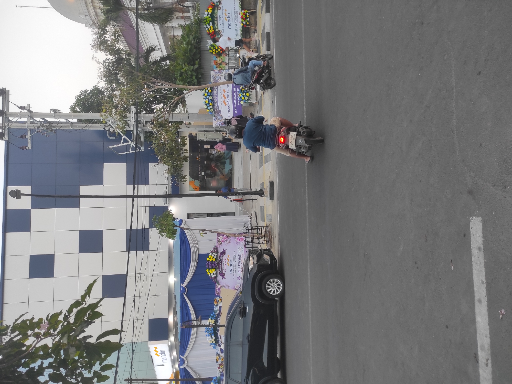 In Bojonegoro, Indonesia, an outdoor scene unfolds with a focus on a bank's new branch opening. The building, identified by the "Mandiri" logo and a checkered facade of white and navy blue, stands prominently. A black SUV is parked in front of the entrance, which is adorned with blue and white drapes and floral arrangements. Two people on motorcycles are visible on the street in the foreground; one rider in a blue shirt is facing away from the camera, while another individual on a separate motorcycle is further down the road. In the background, a white building with a dome is partially visible, suggesting a significant architectural landmark nearby. The sky is overcast, indicating a potential for diffused light or an approaching change in weather. The overall impression is one of a commercial event taking place in a public space.