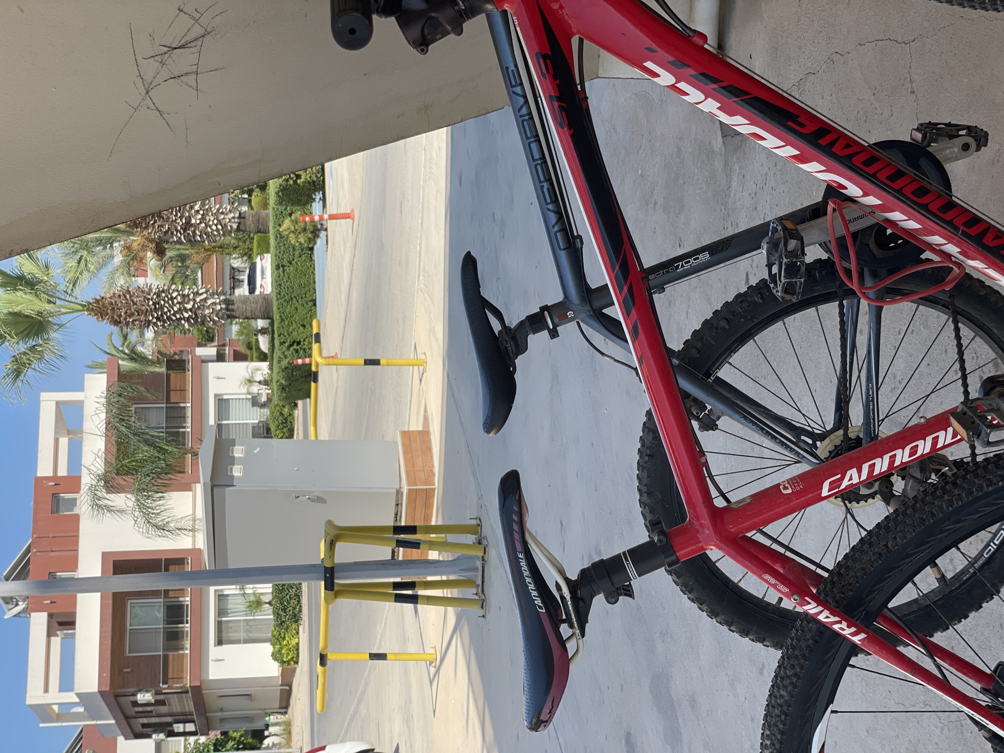 In Trikomo, Northern Cyprus, two bicycles are parked outdoors, leaning against a textured, off-white surface. The bicycle in the foreground is red with black accents and white lettering that reads "CANNONDALE". Its saddle is black with red trim, and the brand "CANNONDALE" is visible on the side. The rear wheel and part of the frame are visible. Behind it, a dark gray bicycle is also parked, with a black saddle that has "CANNONDALE" printed on it. The frame of the gray bicycle has "OVERDRIVE" and "SL3" written on it. In the background, a modern, multi-story building with white and reddish-brown sections is visible, surrounded by greenery and palm trees. Yellow and black barriers and orange traffic cones are also present, suggesting a public or street area. The sky is clear and blue.