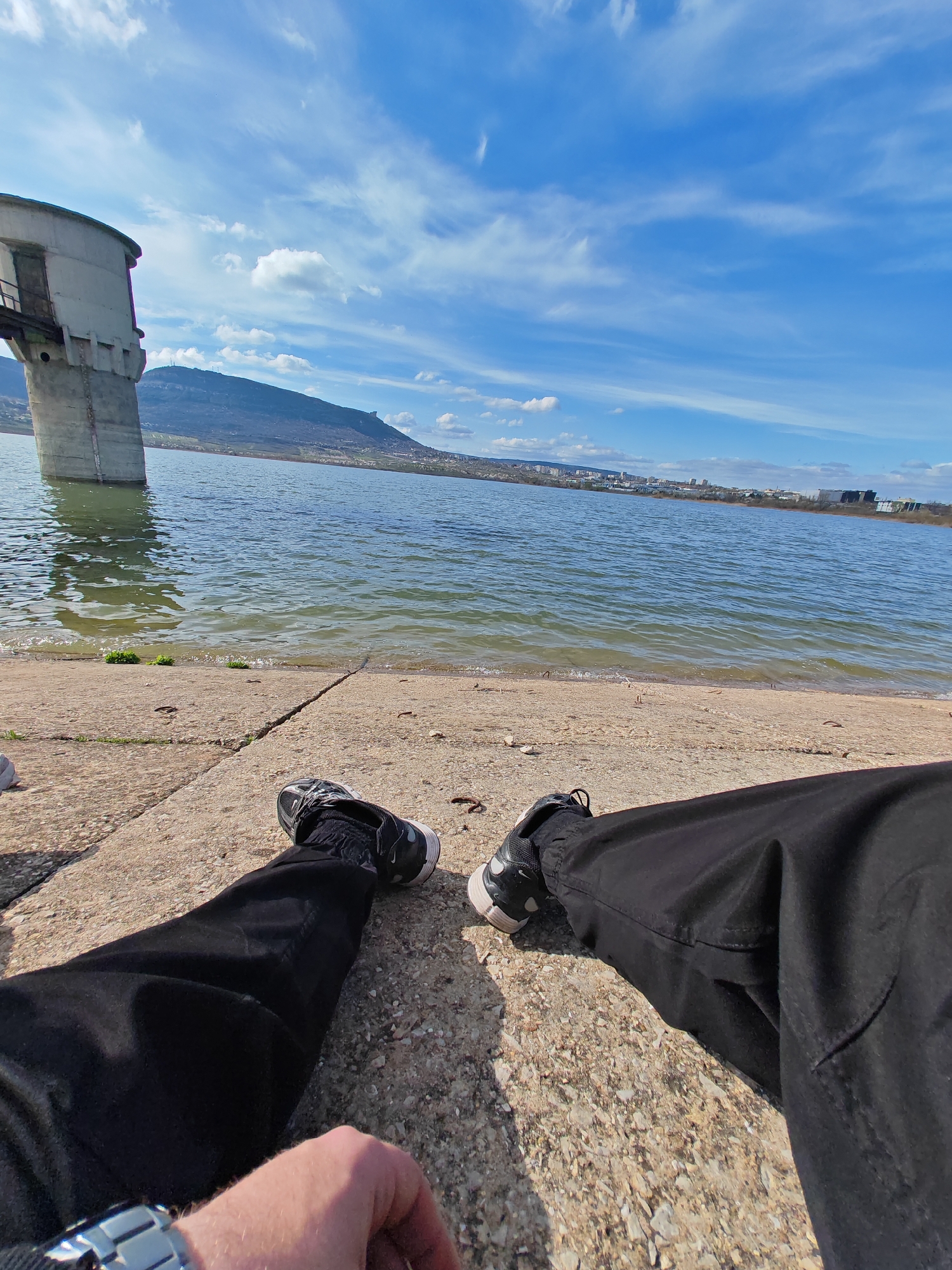 A first-person perspective photograph depicts a person sitting near the shore of a body of water, identified as the Shumen Lake, situated close to the city of Shumen, Bulgaria. The sky is partially cloudy with clear blue patches. The person is wearing black pants and black/white sneakers and is sitting on a concrete surface. In the background, a water tower is partially submerged in the lake, with a mountain range visible further in the distance. Some city buildings are visible near the horizon. The person's left hand with a wristwatch is partially visible in the lower-left corner of the image.