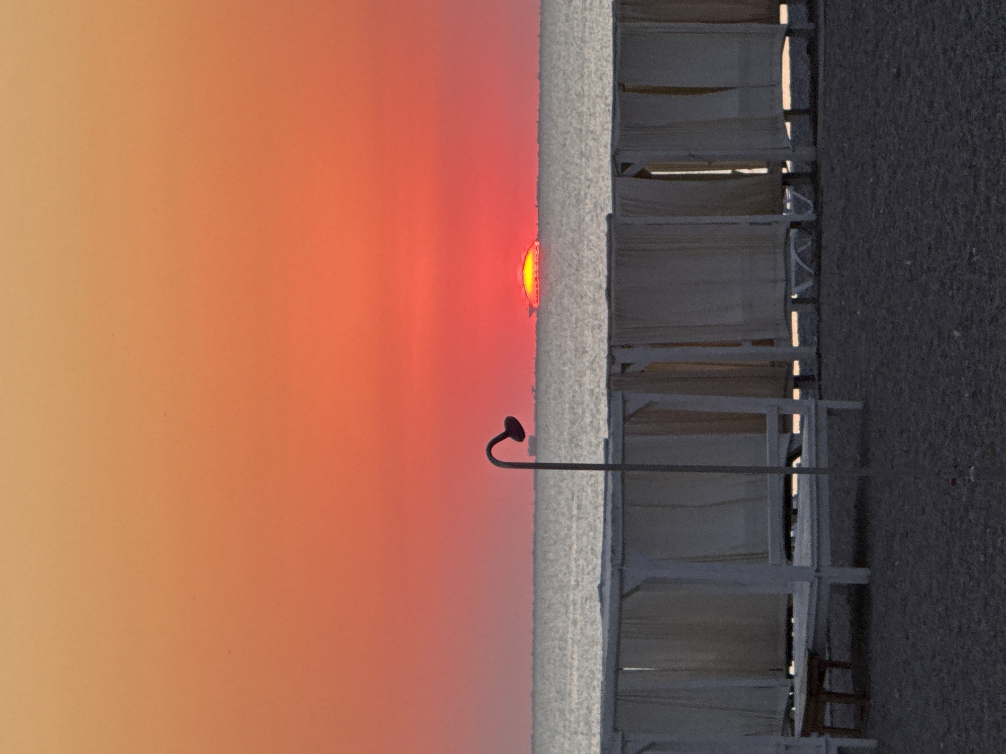 The image depicts a serene sunset scene on a beach in Novofedorovka, Ukraine. The sky is ablaze with vibrant orange and red hues, fading into a softer peach tone towards the top. The sun, a bright red orb with a yellow glow around its lower edge, is partially submerged on the horizon, casting a shimmering path across the calm, grey sea.
In the foreground, several white cabanas with draped beige curtains are lined up on the sandy beach. These structures appear to be beach loungers or private seating areas. A dark, curved lamppost stands to the left of the cabanas, casting a subtle shadow. The beach itself is covered in dark grey pebbles. The overall atmosphere is peaceful and tranquil, characteristic of a coastal sunset.