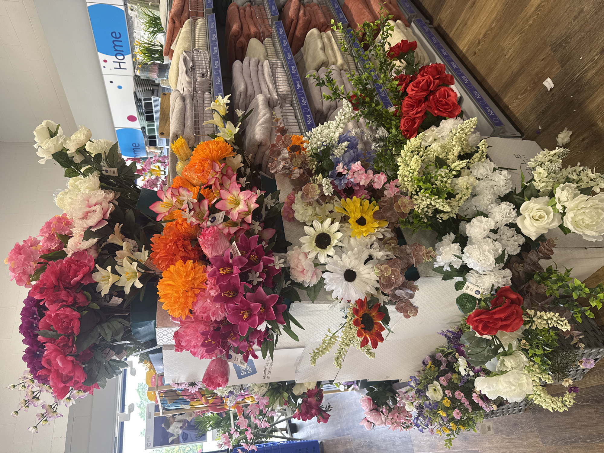 This is a medium shot, angled photograph of a display of artificial flowers in a retail setting. The flowers are arranged in bunches and bouquets, with a wide variety of colors and types, including roses, lilies, sunflowers, and smaller filler flowers. Many of the flowers are vibrant and appear to be made of silk or similar synthetic materials.
In the background, the setting appears to be a store aisle, with shelves displaying folded textiles, possibly towels or blankets, in muted tones of beige, grey, and pink. A blue sign with white text that reads "Home" is visible at the top left of the image, indicating the department or section of the store. Another sign with the word "New" repeated multiple times in a blue strip is visible on the shelves. Some price tags are attached to the flower stems. The floor is a wooden laminate.
The lighting suggests it is daytime, likely indoors with artificial lighting or natural light filtering in from a nearby window or entrance, hinted at by a bright area at the bottom left. There are no people visible in the frame. The overall impression is of a colorful and abundant display of artificial floral arrangements within a retail environment. Visible text includes "Home" and repeated instances of "New." There are also price indications like "£1.29 to £4.99" on a tag.