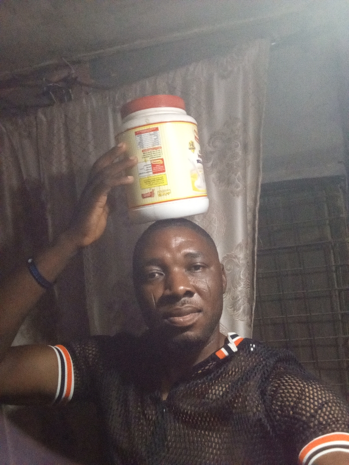 A man balances a large, off-white container with a red lid on his head, holding it in place with his left hand. He wears a black mesh shirt with orange and white stripes on the sleeves and collar. He has short hair, dark skin, and several scars on his face. Background elements include a curtain and what appears to be an air conditioning unit. The lighting is dim.