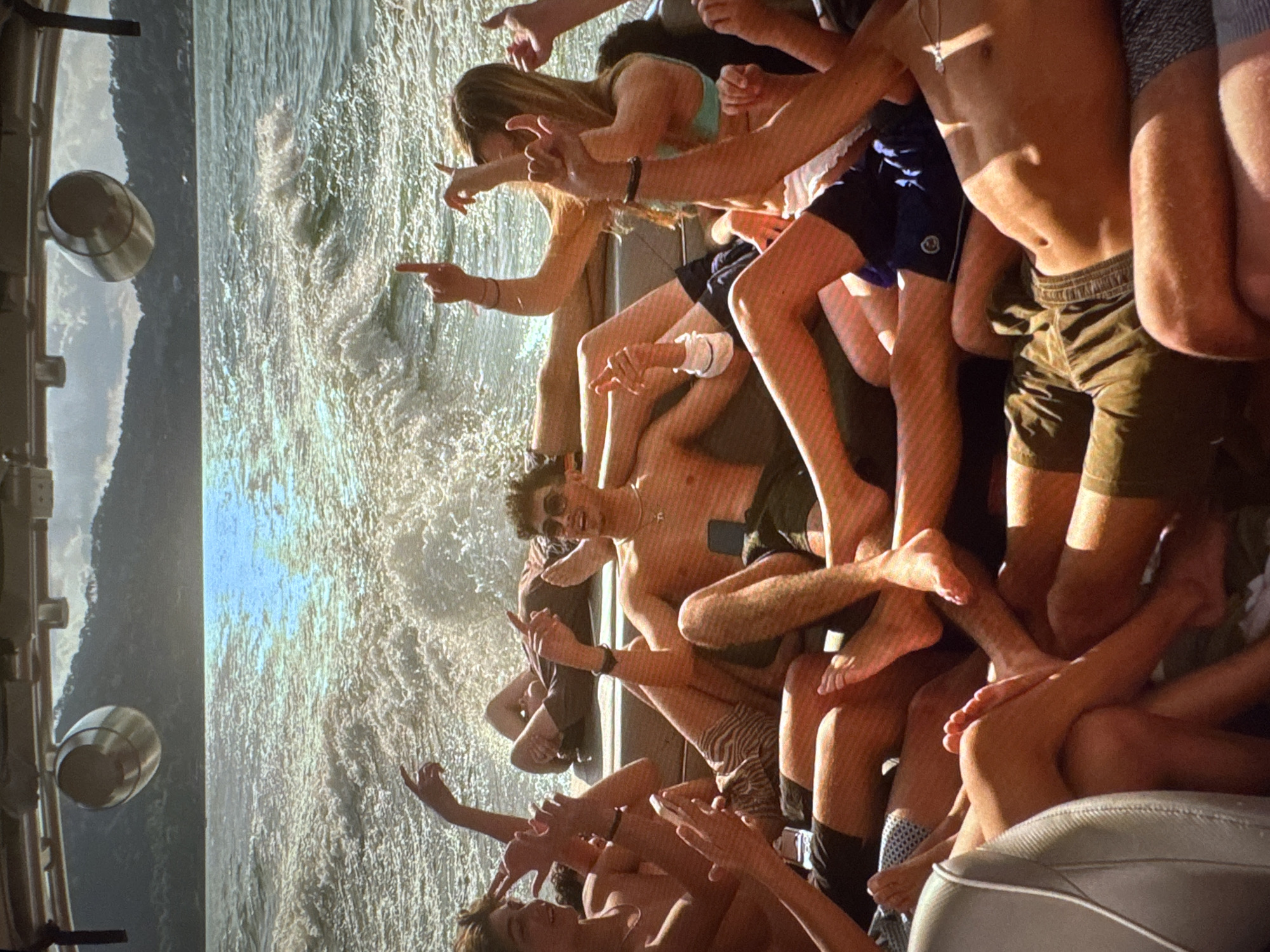 A group of young adults are gathered on the back of a boat, with the sea and mountains in the background. The sun is shining, creating a glittering effect on the water. Several individuals are raising their hands, some making gestures with their fingers. The people are dressed casually, in swimwear and shorts. The scene appears to be a recreational outing on a body of water, possibly a lake or a bay. The location is not in Tallinn, Estonia; the background scenery suggests a warmer climate and different geography.