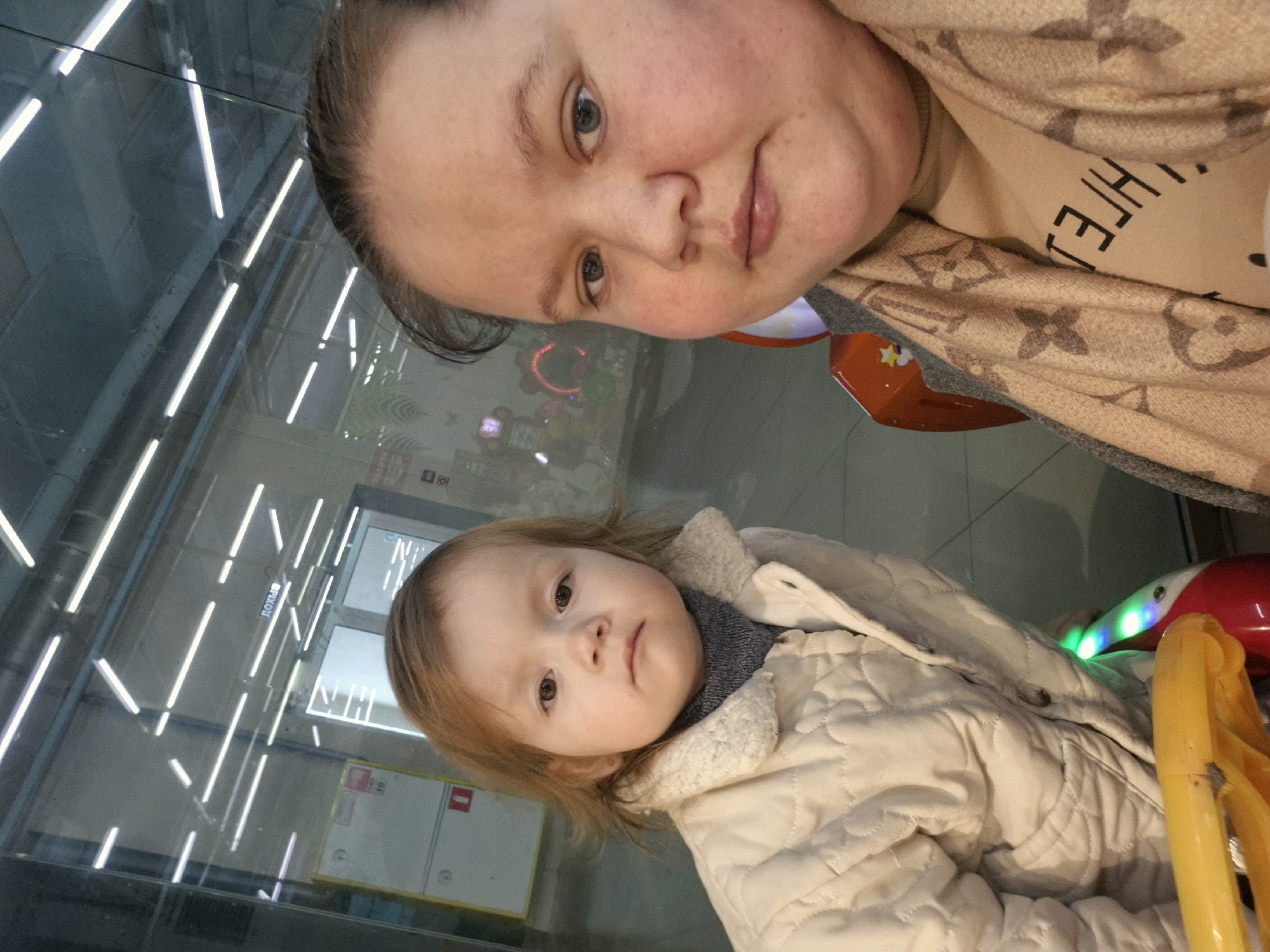 The image shows a woman and a young child indoors. The woman is positioned in the upper portion of the frame, looking down at the camera with a slight smile. She is wearing a tan patterned scarf or garment. The child, who appears to be a toddler, is located in the lower part of the image and is looking directly at the camera. The child is wearing a light-colored, quilted jacket with a furry collar.
The background indicates an indoor environment, potentially a shopping mall or a similar public space, with what seems to be a storefront or a set of doors visible behind the child. The reflection on the glass suggests the presence of fluorescent lighting. Part of a ride or toy is seen on the right side of the frame, with yellow and green lights, which may imply an entertainment area. The location is specified as Unknown, Unknown.