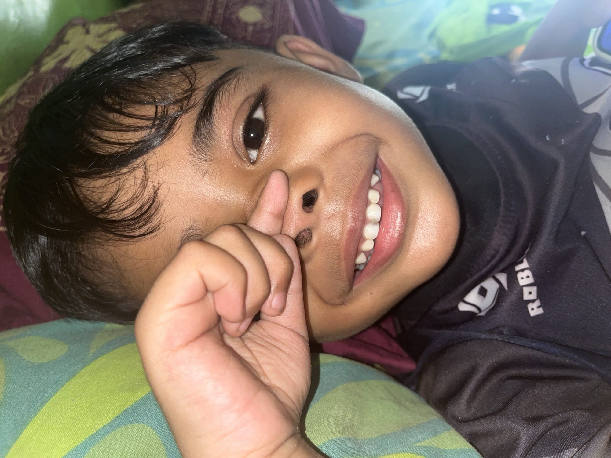 The image shows a close-up, eye-level shot of a young boy lying down and looking at the camera. He is resting on a patterned surface, likely a bed or couch, with a dark patterned cloth underneath his head and a green and yellow patterned cloth visible on the left side of the frame. The boy has dark hair, dark eyes, and is smiling, revealing his teeth. His right hand is raised, with his index finger touching his nose. He is wearing a black t-shirt with a white logo that appears to be the "Roblox" logo. The lighting suggests an indoor setting, possibly during the day with natural light contributing to the illumination. The scene is devoid of any specific urban landmarks that would allow for precise location within Bumi Mulyo, Indonesia, beyond the general context of being a residential interior.