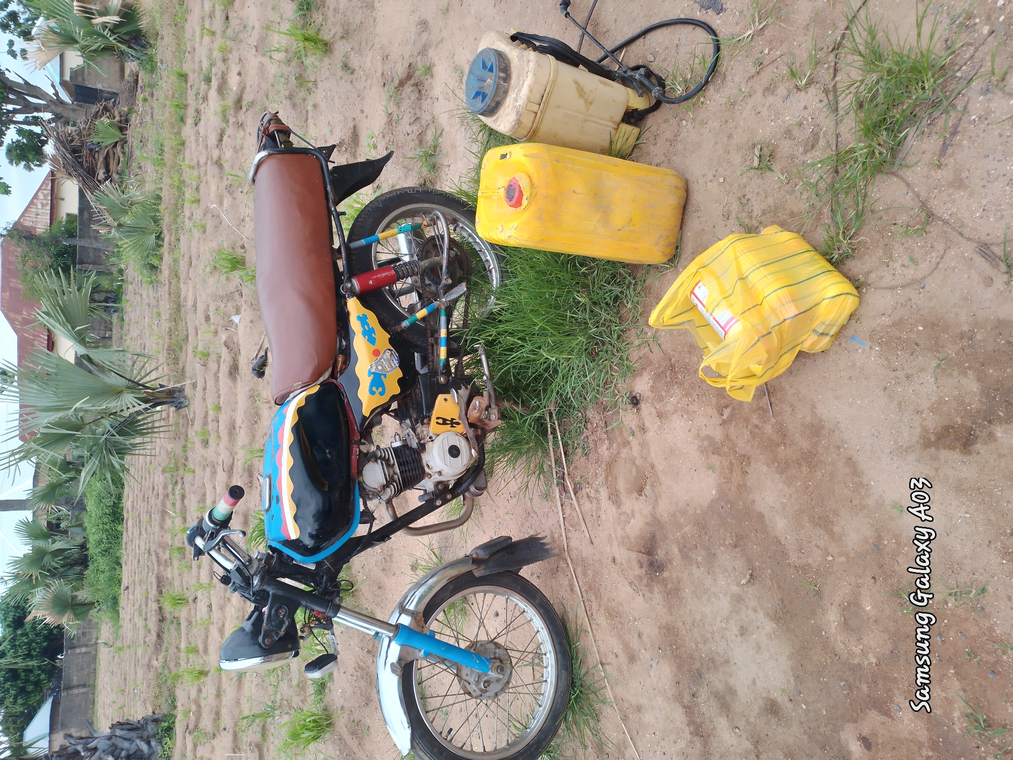 The image depicts an outdoor scene in Angwan Hausawa, Nigeria. A motorcycle, painted with black, blue, yellow, and red stripes on its fuel tank, is parked on a dirt ground. The motorcycle has a brown seat and a chrome front fender. To the right of the motorcycle are two yellow plastic containers and a yellow plastic bag. One of the containers is tall and cylindrical with a red cap, while the other is shorter and rectangular with a blue lid. The yellow plastic bag, filled with items, is positioned in the foreground. The environment consists of dry, sandy soil with patches of green grass. In the background, palm trees and some buildings with red roofs are visible. A watermark at the bottom left corner reads "Samsung Galaxy A03".