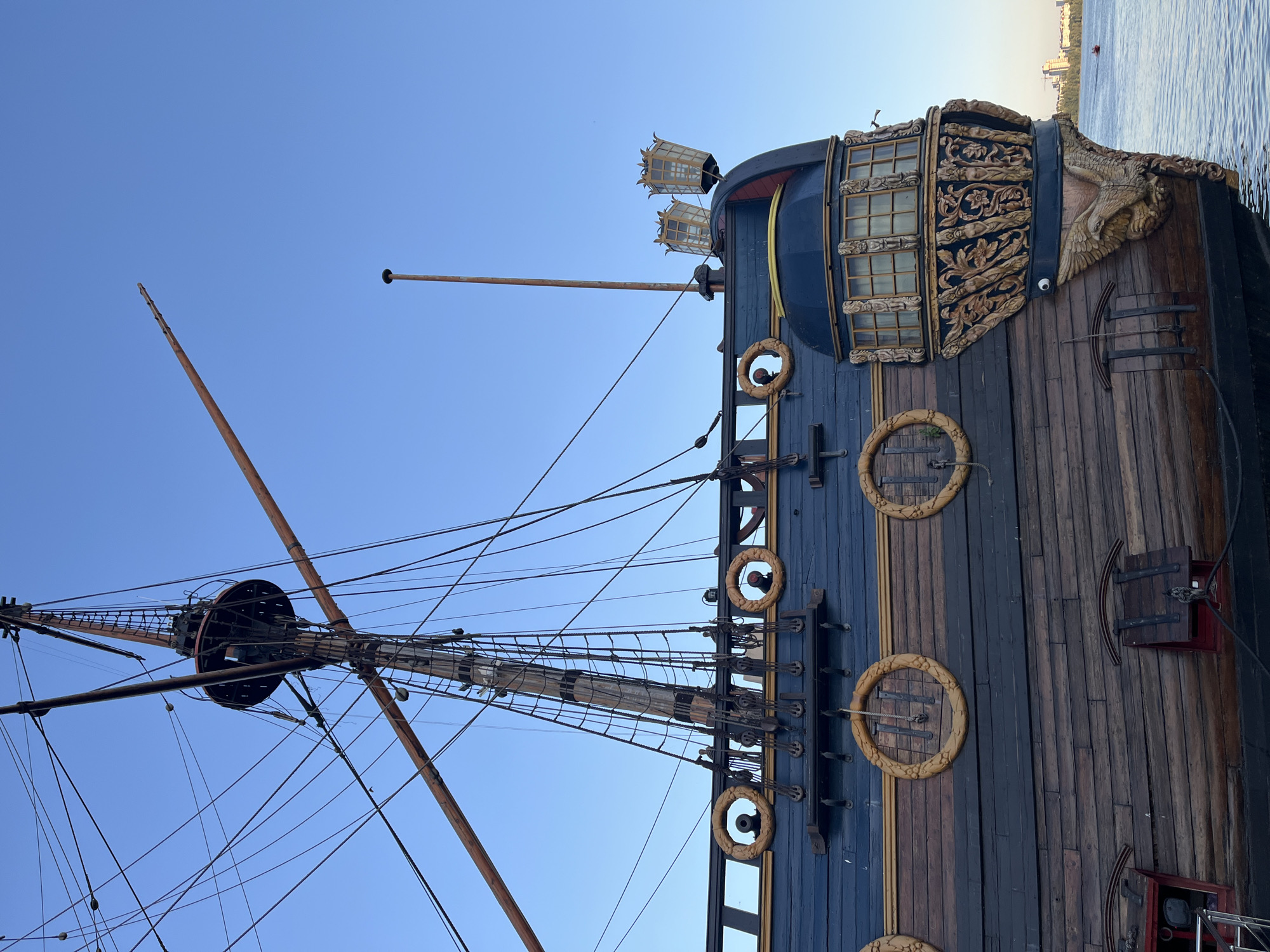 The image shows the stern of a historic wooden sailing ship docked on a body of water. The ship has dark blue planking on its upper deck, adorned with ornate golden circular decorations and what appear to be cannons peeking through them. The lower hull is made of weathered dark wood planks. At the stern, there is a beautifully carved wooden eagle figurehead, with intricate gold detailing and carvings of figures and floral motifs above it. Two ornate lamps with golden frames and glass panels are visible on the deck railing. The ship's mast and rigging are prominent in the background, set against a clear blue sky. In the distance, across the water, a city skyline with buildings and a bridge is visible. A small red boat is also present on the water. This scene is located in the city of Novaya Usman, Russia.