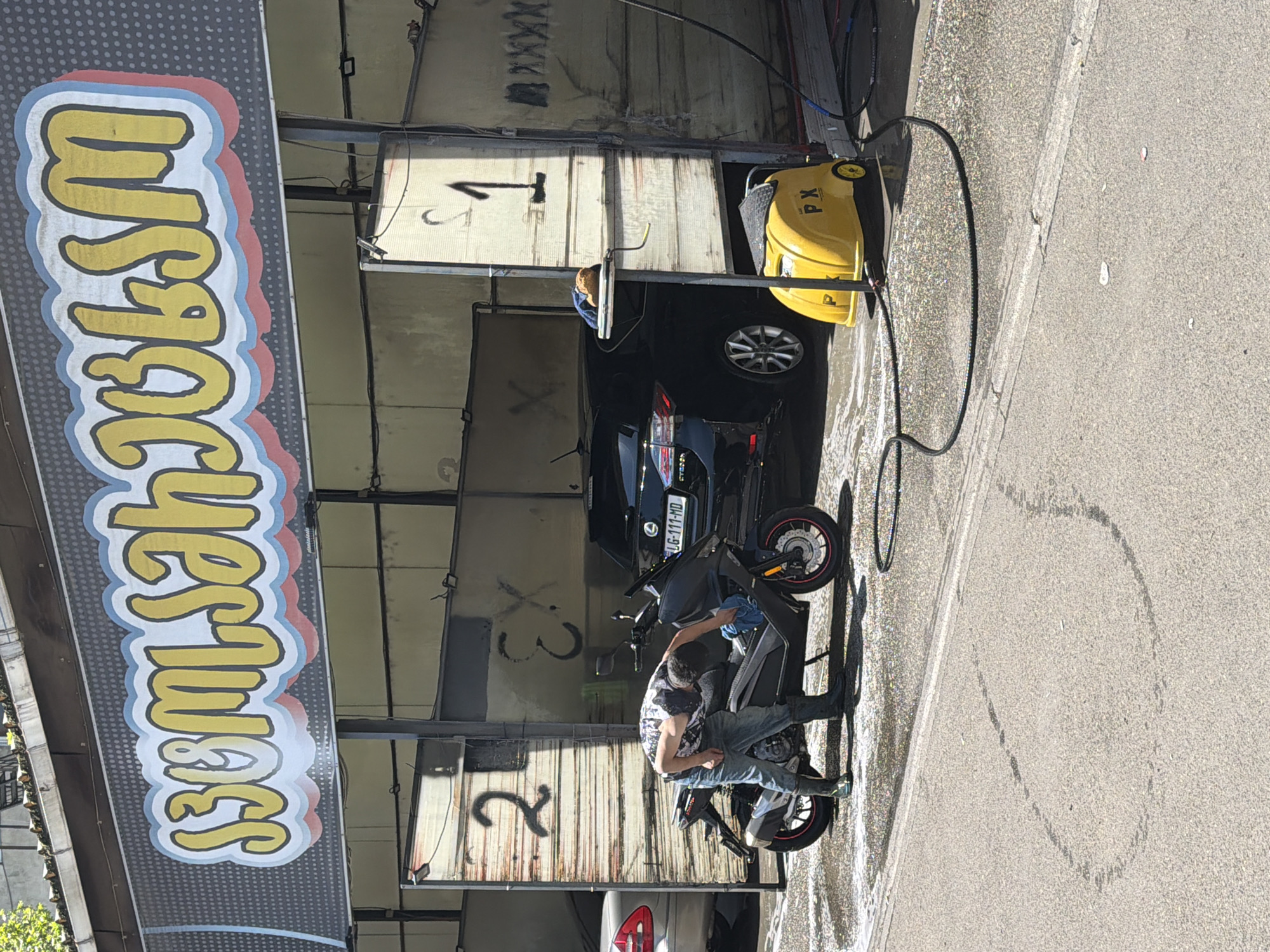 A man is washing a black scooter in a car wash bay. The scooter is parked in the second bay of a three-bay car wash. A black sedan is parked in the first bay. There is a yellow pressure washer beside the car. The car wash is located in Tbilisi, Georgia, as evidenced by the license plate on the black sedan, which reads "LG-111-MD." The sign above the car wash reads "Wash."
