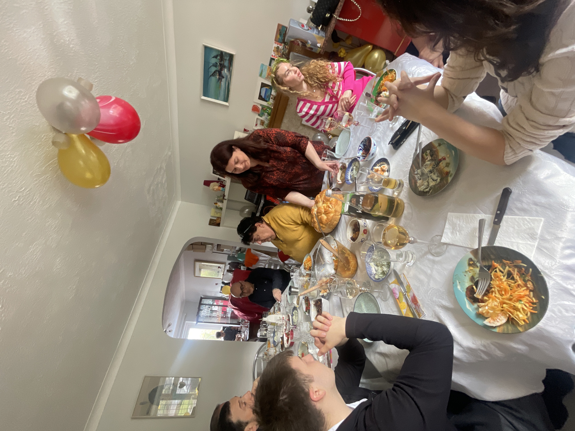 The image depicts a gathering of people around a table laden with food and drinks. The location appears to be indoors, potentially a dining room, evidenced by the presence of a long table covered with a white tablecloth, various dishes and glassware, and multiple people seated around it.
The individuals in the image are diverse in age and gender. They are engaged in eating, drinking, and conversation, suggesting a social occasion, likely a meal or celebration.
Several objects are visible on the table, including plates of food, glasses, bottles of beverages (including wine), and cutlery. Other objects are scattered around the room, including balloons attached to the wall.
While the exact location within Maidstone, United Kingdom is not discernible from the image, the setting is indicative of a domestic environment in a residential area.