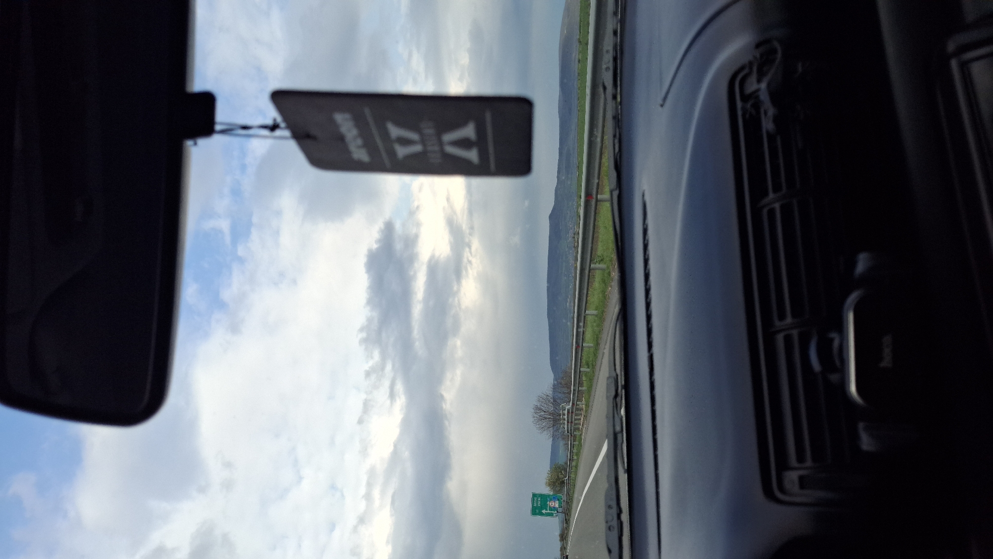 The image is taken from inside a car, looking out the front windshield. A rear-view mirror and an air freshener hang from the top of the windshield. The outside scene features a cloudy sky and a road with green vegetation and a distant mountain range on the right. A green road sign is visible on the left side of the road. The road appears to be a highway. Based on the road sign "Златина 29 км" (Zlatina 29 km), the car is located approximately 29 kilometers away from the city of Zlatina, Bulgaria. The car's dashboard and air vents are visible on the right side of the frame.