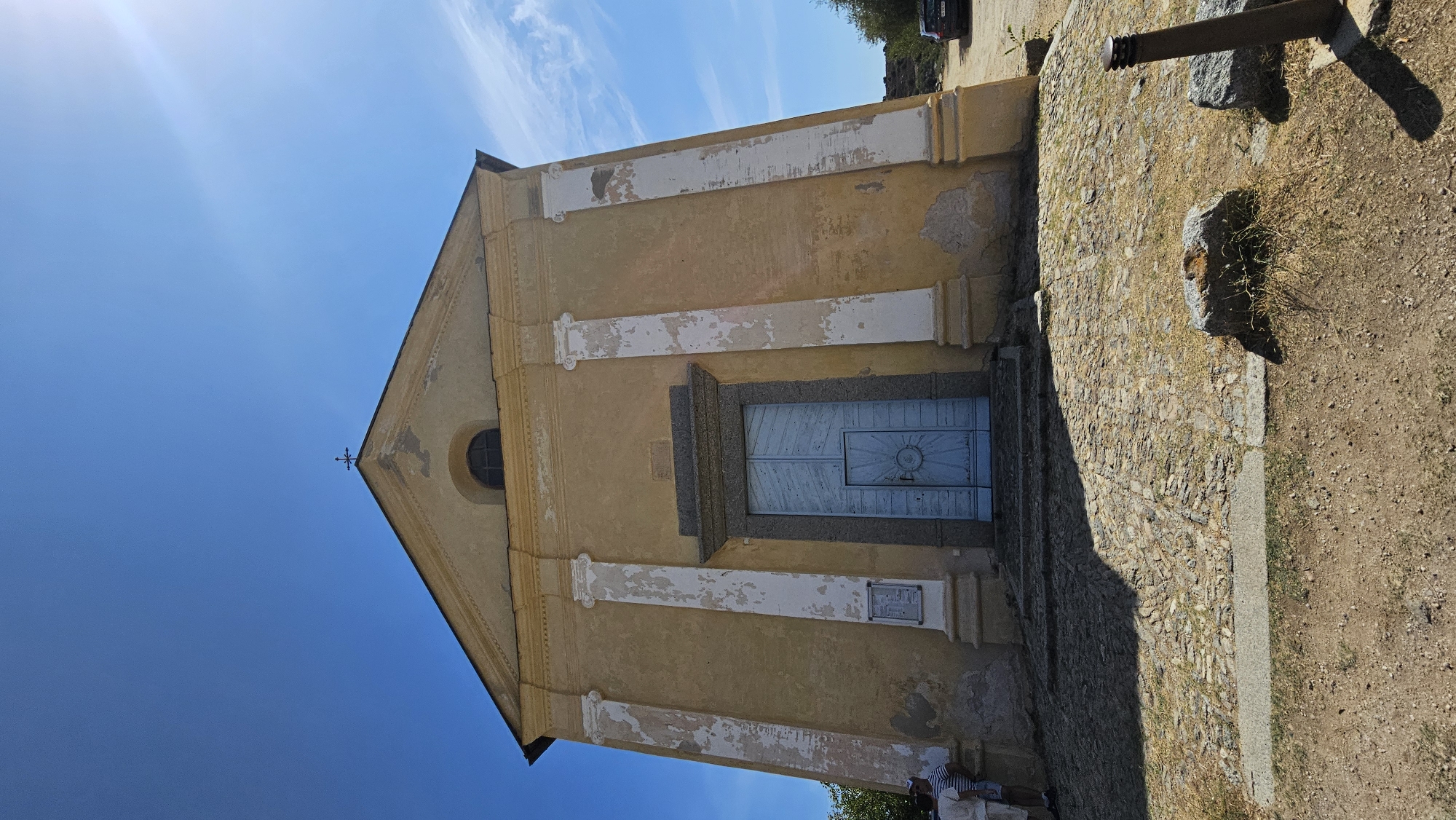 The image shows a low-angle, wide shot of the facade of a weathered, mustard-yellow church with a pale blue double door. The church has a triangular pediment at the top, topped with a small cross. Four fluted pilasters, painted white and showing significant peeling, flank the entrance. Above the doors, a small, arched window is visible, dark with age or grime. A plaque is mounted on the left side of the facade, near the ground. The ground in front of the church is paved with irregular cobblestones, interspersed with dry grass and dirt. Two individuals, dressed in light-colored clothing, are standing to the left of the entrance, looking towards the church. In the background, to the right, a dark-colored car is parked near some trees. The sky above is a clear, bright blue with wispy white clouds. The overall impression is of an old, somewhat neglected religious building situated in a dry, possibly rural environment. This church is located in the village of Sant'Antonino, Corsica, France.