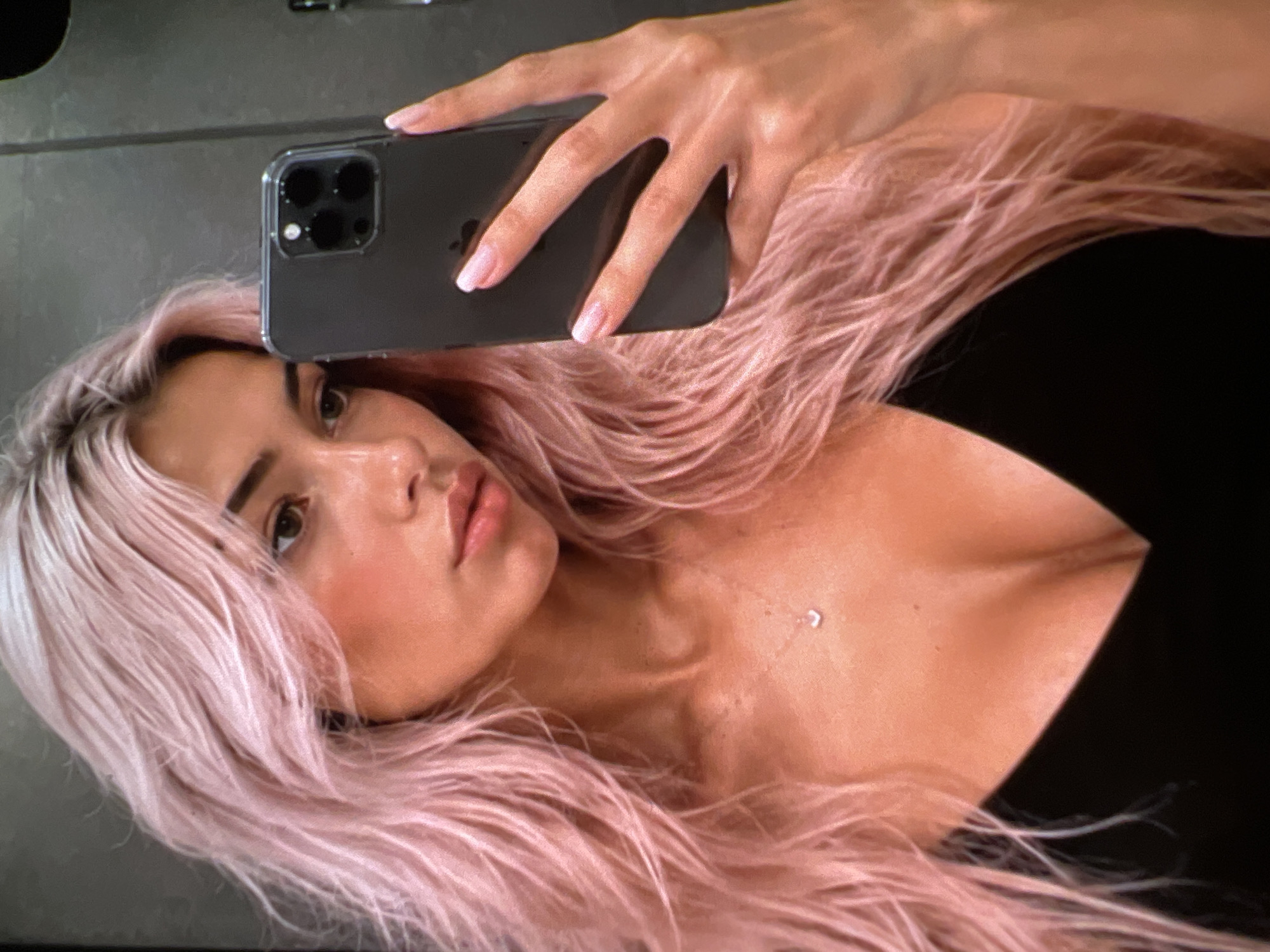 A young woman with long, pink, wavy hair is taking a selfie in a mirror. She is wearing a black top and a small heart-shaped pendant necklace. Her right hand is holding a dark-colored smartphone. The background appears to be indoors, possibly a bathroom or dressing area, with a gray, textured wall. The lighting is warm and suggests an indoor setting.