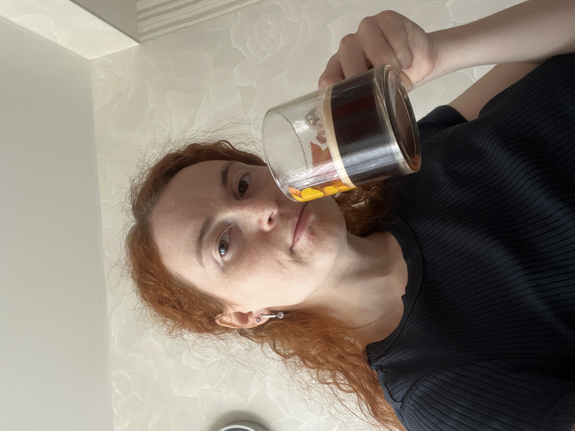 A red-haired woman with freckles is holding a clear glass mug filled with a dark liquid and some orange slices, possibly tea. She's wearing a black ribbed shirt and a silver ear cuff earring. The background is a white wall with a subtle floral pattern. Based on the location metadata, the photo was taken in the city of Chaikovsky, Russia.