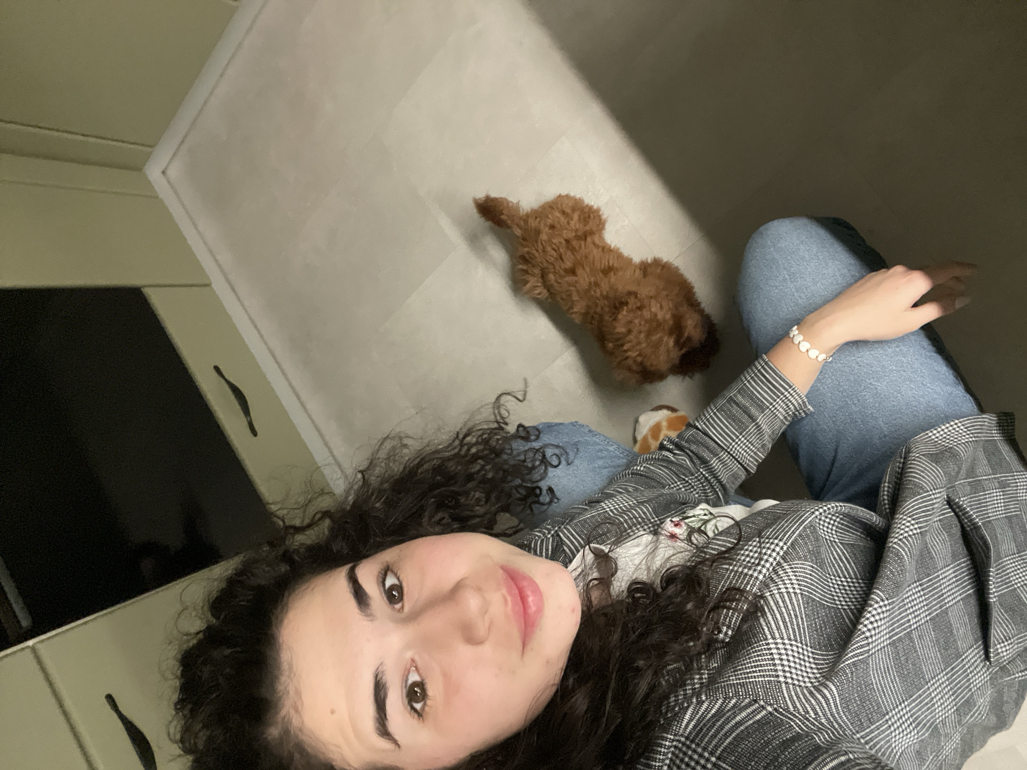 A woman with curly dark hair and wearing a gray plaid blazer and blue jeans sits on the floor, angled towards the camera. A small brown poodle dog is lying on the floor behind her. The floor is a light gray tile. A cabinet with black handles is visible in the background, and a white wall is behind it. The image was most likely taken indoors. It is not possible to determine if the photo was taken in the city of Sofia, Bulgaria with the provided information.