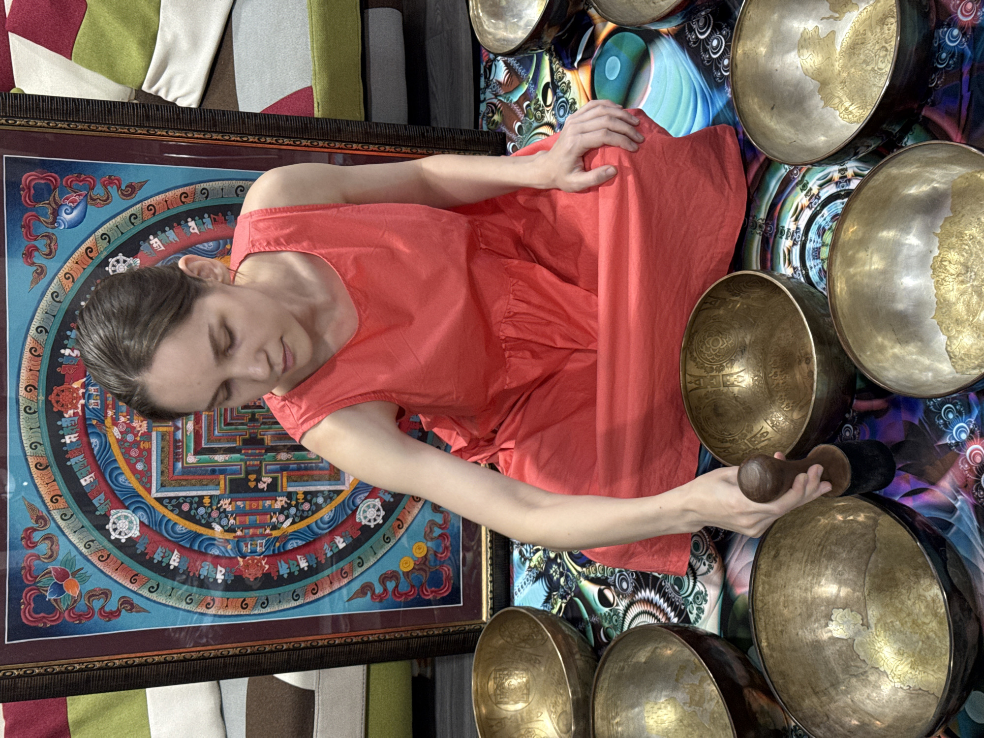 A woman in a coral dress sits next to a framed mandala art piece. She is reaching out with a wooden mallet toward a collection of singing bowls. The background includes patterned fabrics and a psychedelic-looking backdrop. The specific location in Tashkent, Uzbekistan, is not evident from the image.