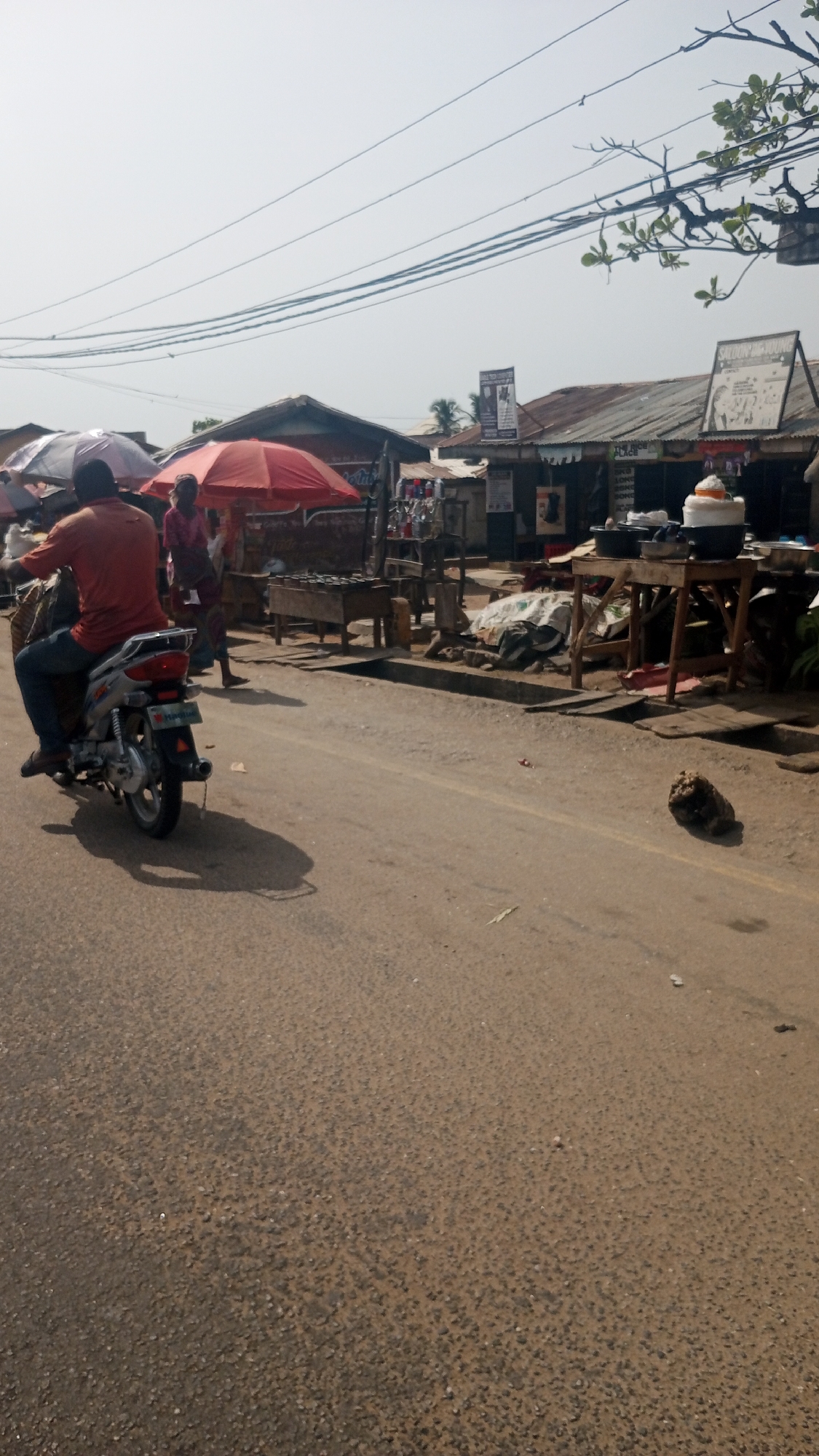 An outdoor scene in Lokoja, Nigeria, depicts a street with various activities and structures. A man is riding a motorcycle on the left side of the road. Adjacent to the road is a row of businesses with red and grey umbrellas providing shade. Several people are standing or walking near these businesses. The structures vary, some appearing to be shops with metal roofs, and signage. Power lines run overhead. The road surface is worn. A rock or similar object lies on the street.