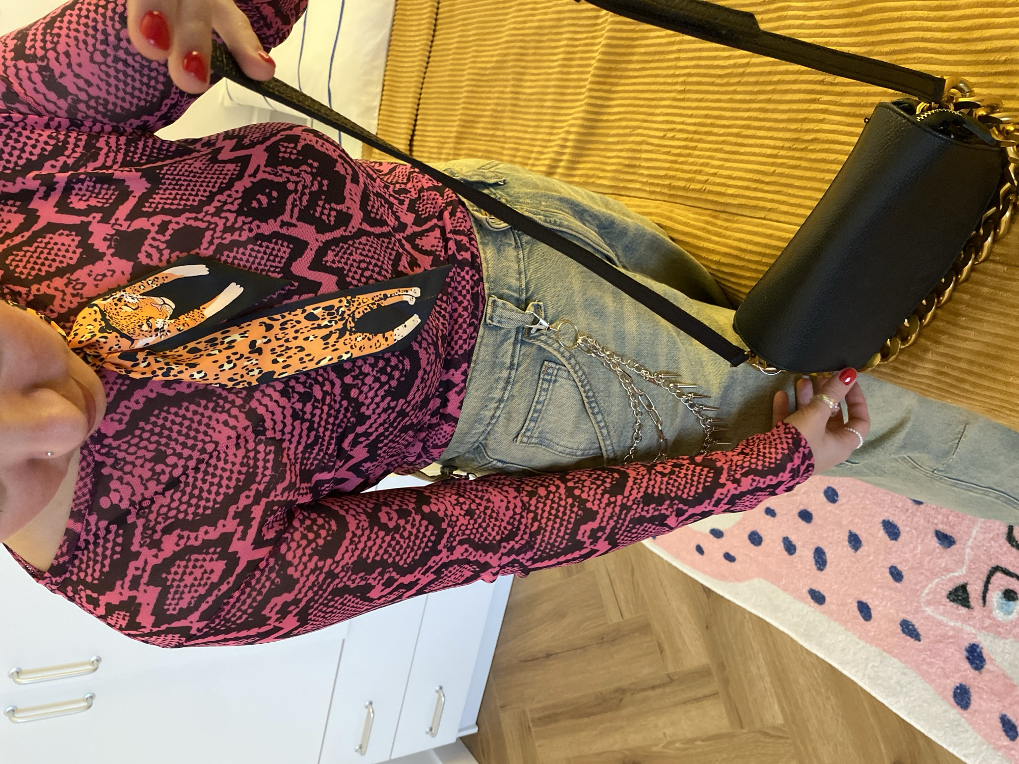A person is standing in a room, likely a bedroom, wearing a pink and black snake-print long-sleeved top, light-wash jeans with a chain accessory, and a black crossbody bag with a gold chain strap. The person has red nail polish. The background includes a white dresser with silver handles, a wooden floor, a pink rug with blue polka dots and a cartoon cat design, and a yellow corduroy textured surface, possibly a bed. The image quality is slightly grainy. There is no identifiable information suggesting the location is Izhevsk, Russia.
