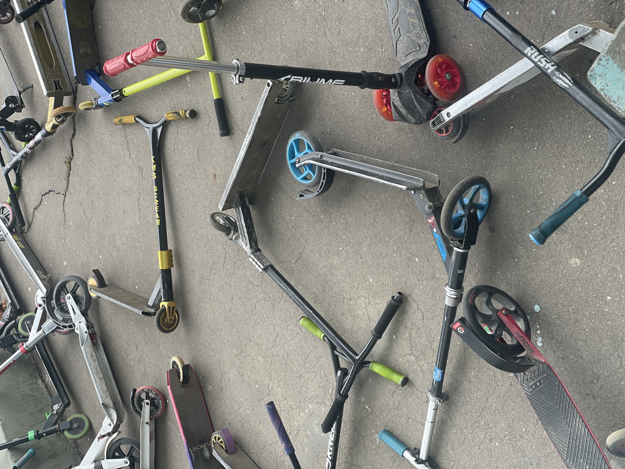 The image shows a collection of scooters lying on a concrete surface, possibly a sidewalk or paved area. The scooters are of various designs and colors, including silver, black, red, blue, yellow, and green. Some appear to be children's scooters, while others may be adult models. The scooters are scattered in a disorganized manner.
The location in the city of Moscow, Russia cannot be determined from this image. There is no indication of the environment that would give the indication of the city, such as buildings, signs, architectural landmarks, or other contextual elements.