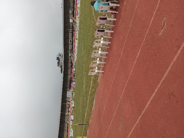 A stadium scene under an overcast sky features a red running track marked with white lines. A row of white plastic chairs sits along the track's edge, bordering a green field. In the background, a stadium structure is visible, adorned with sponsor banners. A small group of people is gathered in the stands.