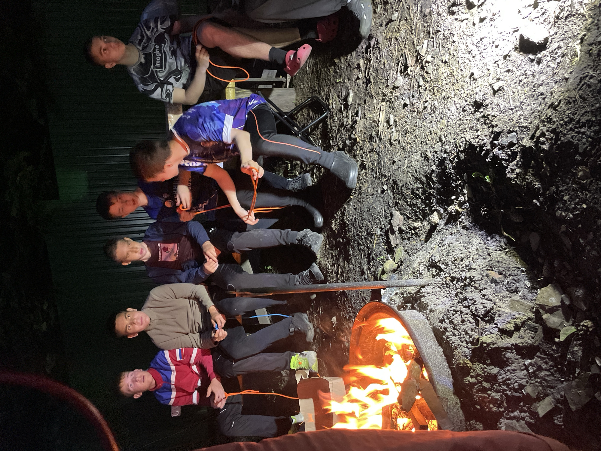 In Nikolaevka, Russia, six young males are seated outdoors at night around a bonfire. The bonfire is contained within a metal ring on the ground, which is covered in dark soil and small stones. The males are seated on a wooden bench and folding chairs, facing the fire. They are dressed in casual clothing, including t-shirts, jackets, and pants. Several of the males are holding orange ropes, and their expressions suggest they are engaged in an activity or conversation. The background is dark and appears to be a natural setting, possibly a backyard or campsite, with a dark fence or wall visible. The lighting is primarily from the bonfire, casting a warm glow on the scene and creating shadows.