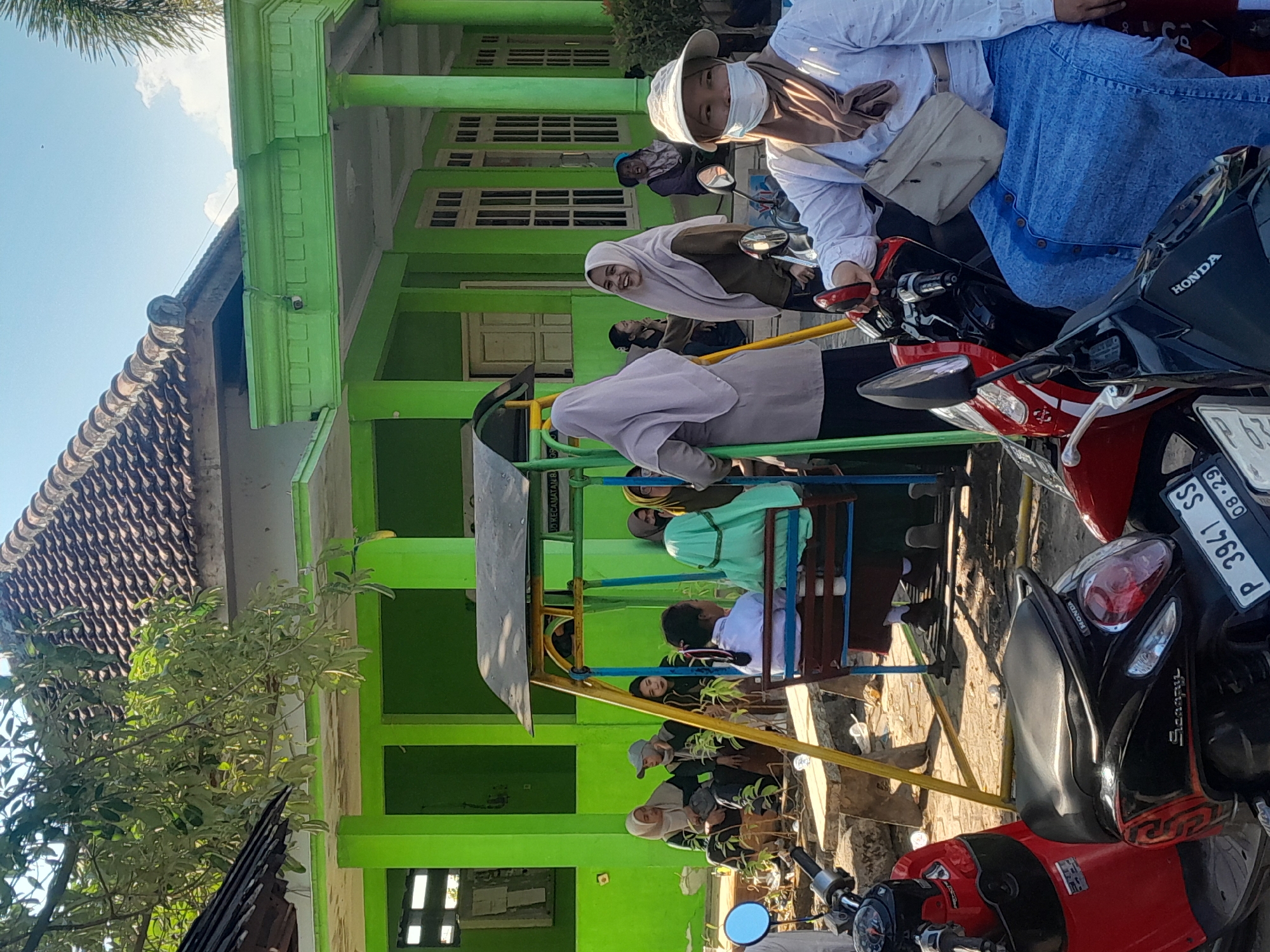 In Sambirejo, Indonesia, on a sunny day, a group of people are gathered outside a bright green building with a brown tiled roof. The building appears to be a school or community center, with windows lining the facade and pillars supporting an overhang. A small playground structure with a swing set is present in the foreground. Several individuals, mostly women wearing hijabs and modest clothing, are interacting and seated on the ground and near the playground. Some are looking towards the playground, while others are engaged in conversation. In the immediate foreground, two motorcycles are parked, one red and one black. A woman wearing a white shirt, a light-colored hat, and a face mask is seated on the black motorcycle. The scene depicts a casual gathering in an outdoor setting, possibly after school or during a community event.