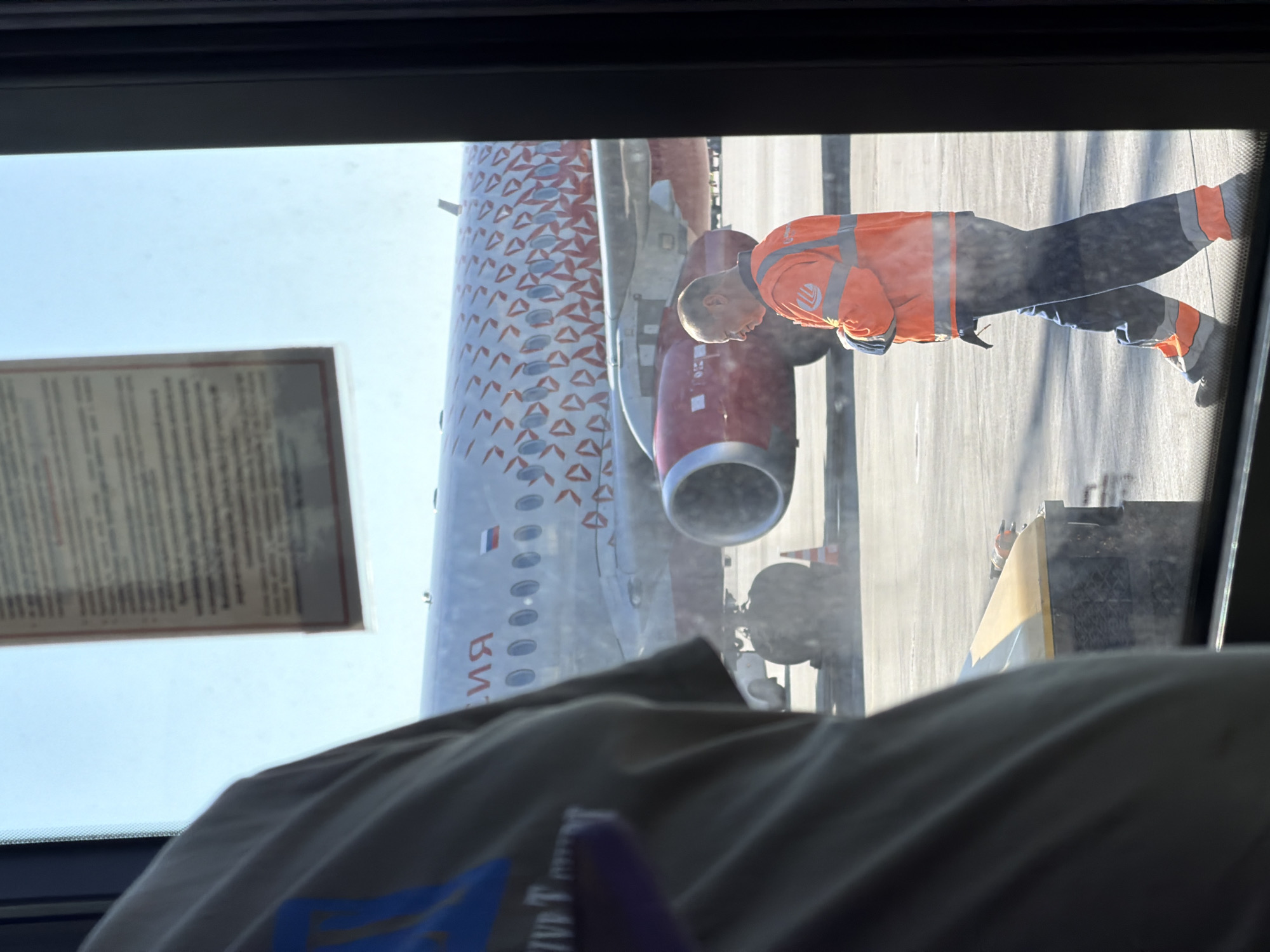 An airport worker in an orange high-visibility jacket and blue pants with reflective orange stripes walks across the tarmac near an airplane at Khimki's Sheremetyevo International Airport. The airplane, with a distinctive red and white patterned fuselage, is parked with its engines visible. In the foreground, partially obscuring the view through a window, is the back of a person wearing a light-colored shirt with blue lettering. A document with text is taped to the window above the airplane. The scene is illuminated by bright daylight, casting shadows on the ground.
