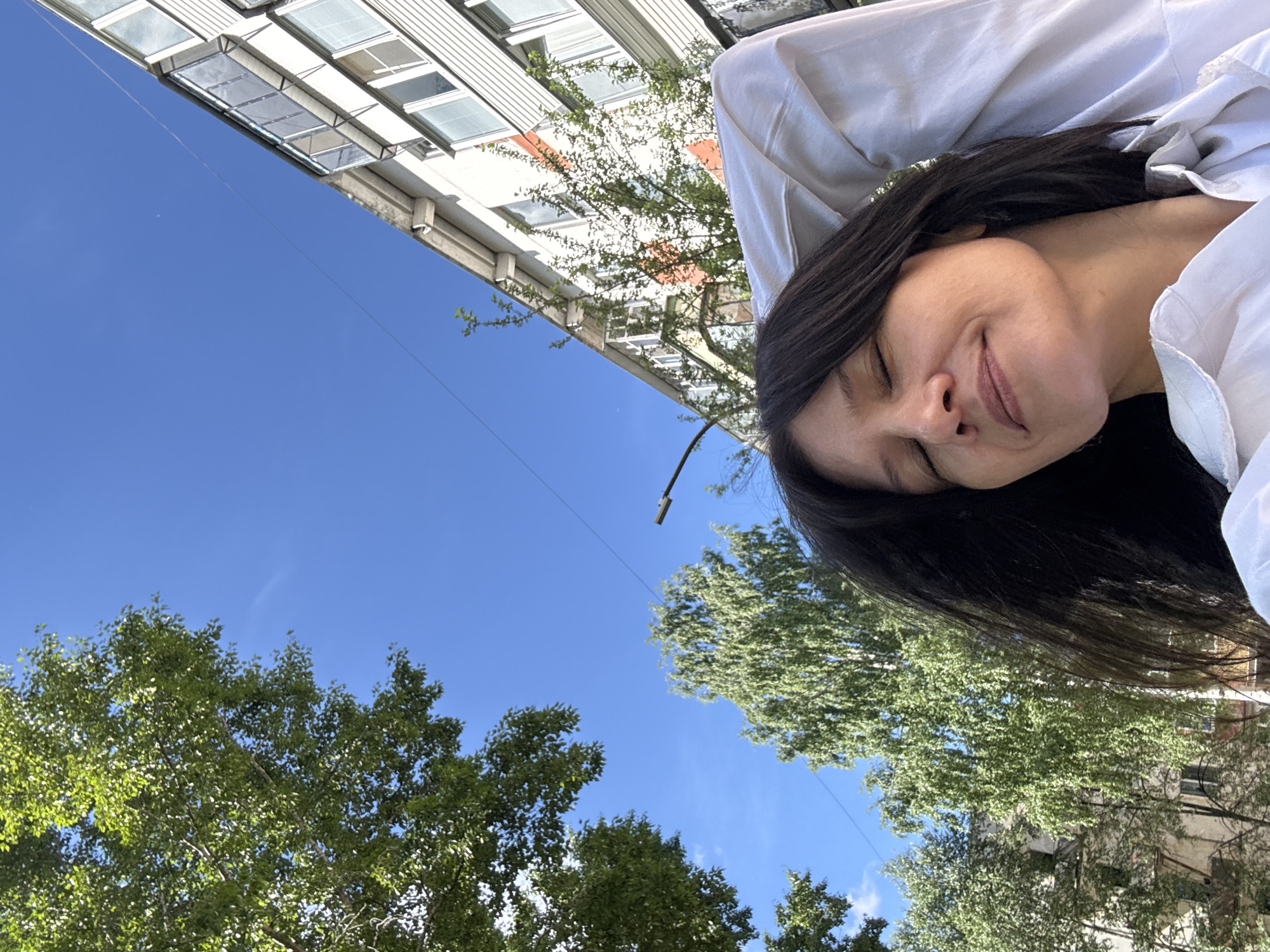 The image shows a woman from the shoulders up, with her eyes closed and a slight smile on her face. She is wearing a white shirt and her arms are raised behind her head. The background consists of a bright blue sky, green trees, and a multi-story building. The perspective suggests the photo was taken outdoors during the day. The building appears to be an apartment block, typical of urban areas. Based on the visual cues, the scene is likely set in the city of Chaikovsky, Russia.