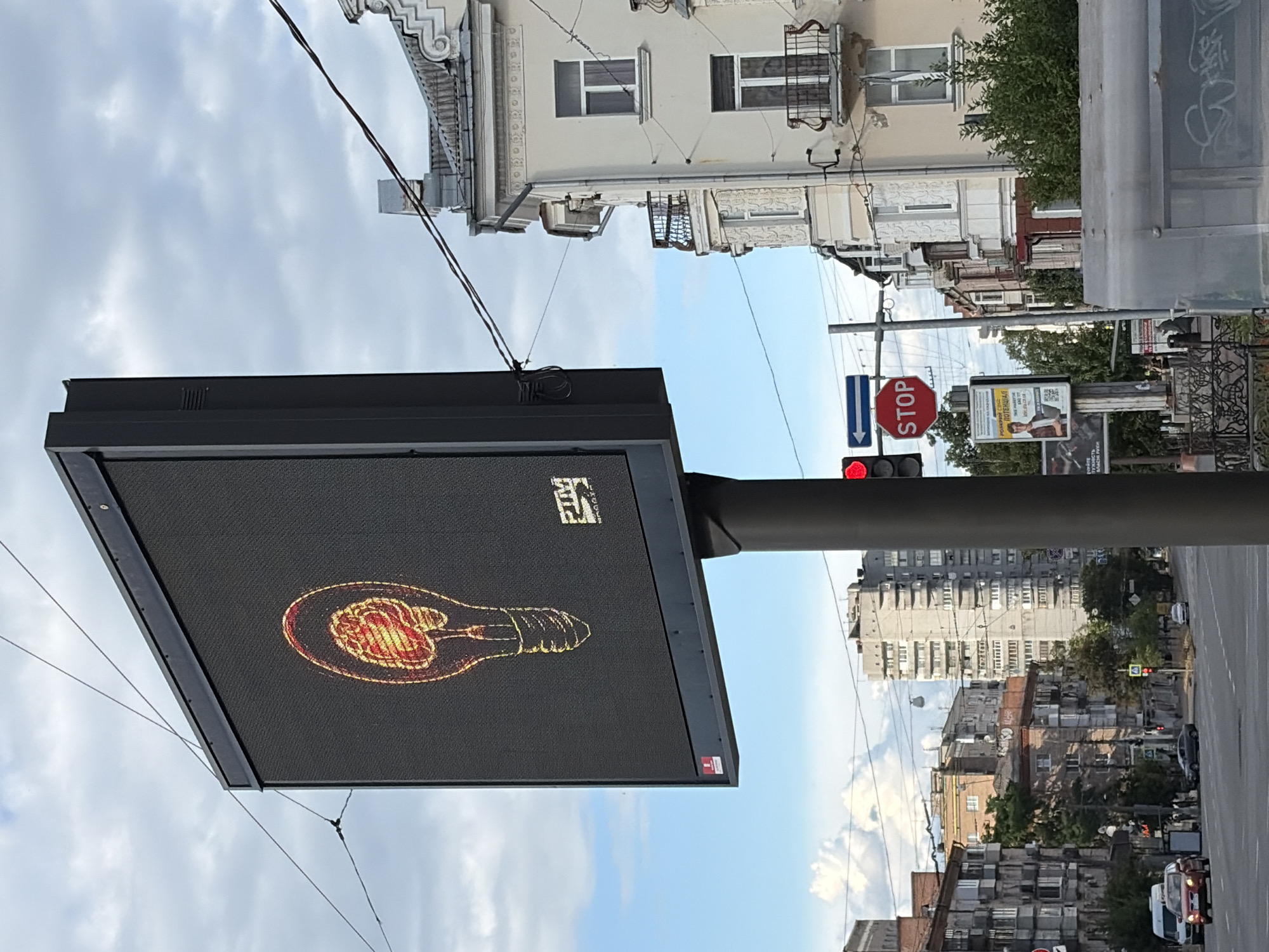 The image depicts a street scene in Chernihiv, Ukraine, with a prominent digital billboard displaying a lightbulb with a human brain inside, rendered in yellow and red hues. The billboard is mounted on a dark gray pole. To the right of the billboard, a red stop sign is visible, accompanied by a blue arrow sign indicating a left turn. Several buildings line the street, varying in architectural style and height, with some featuring balconies. Power lines crisscross the sky. A few vehicles are present on the road, including a red car in the foreground and a white van. The sky is partly cloudy, with patches of blue sky visible. The overall impression is of a typical urban environment.